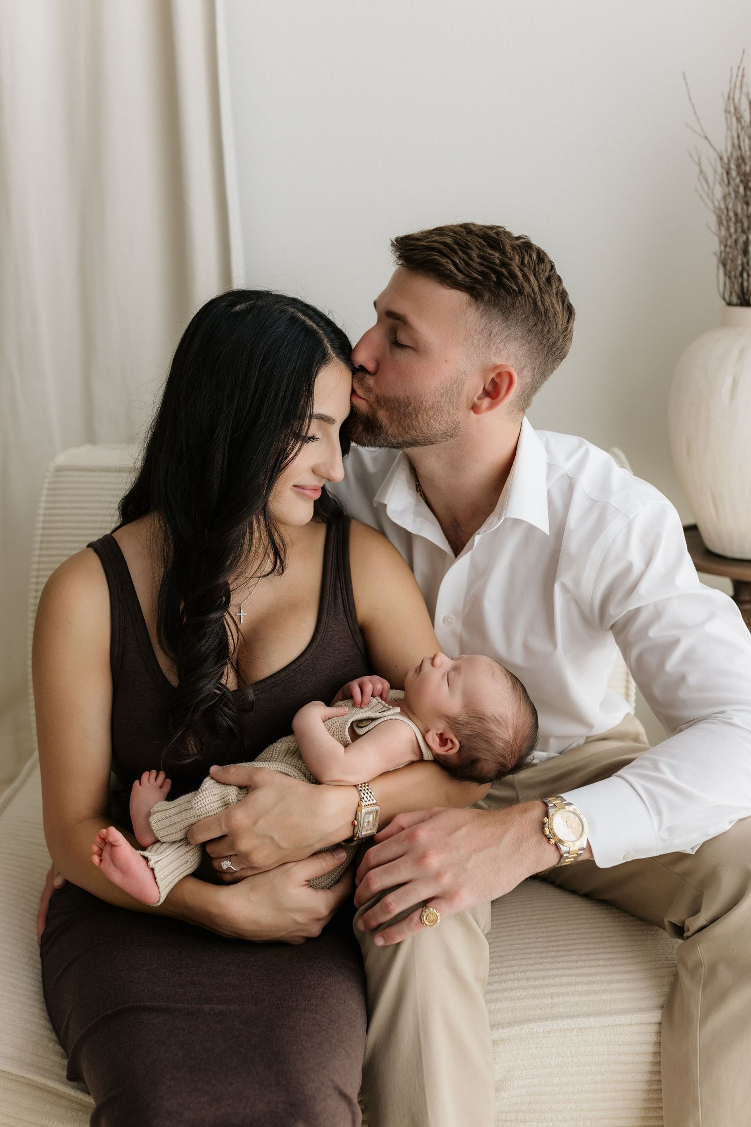 A couple shares a tender kiss while cradling their newborn baby, seated together in a softly lit, neutral-toned room.