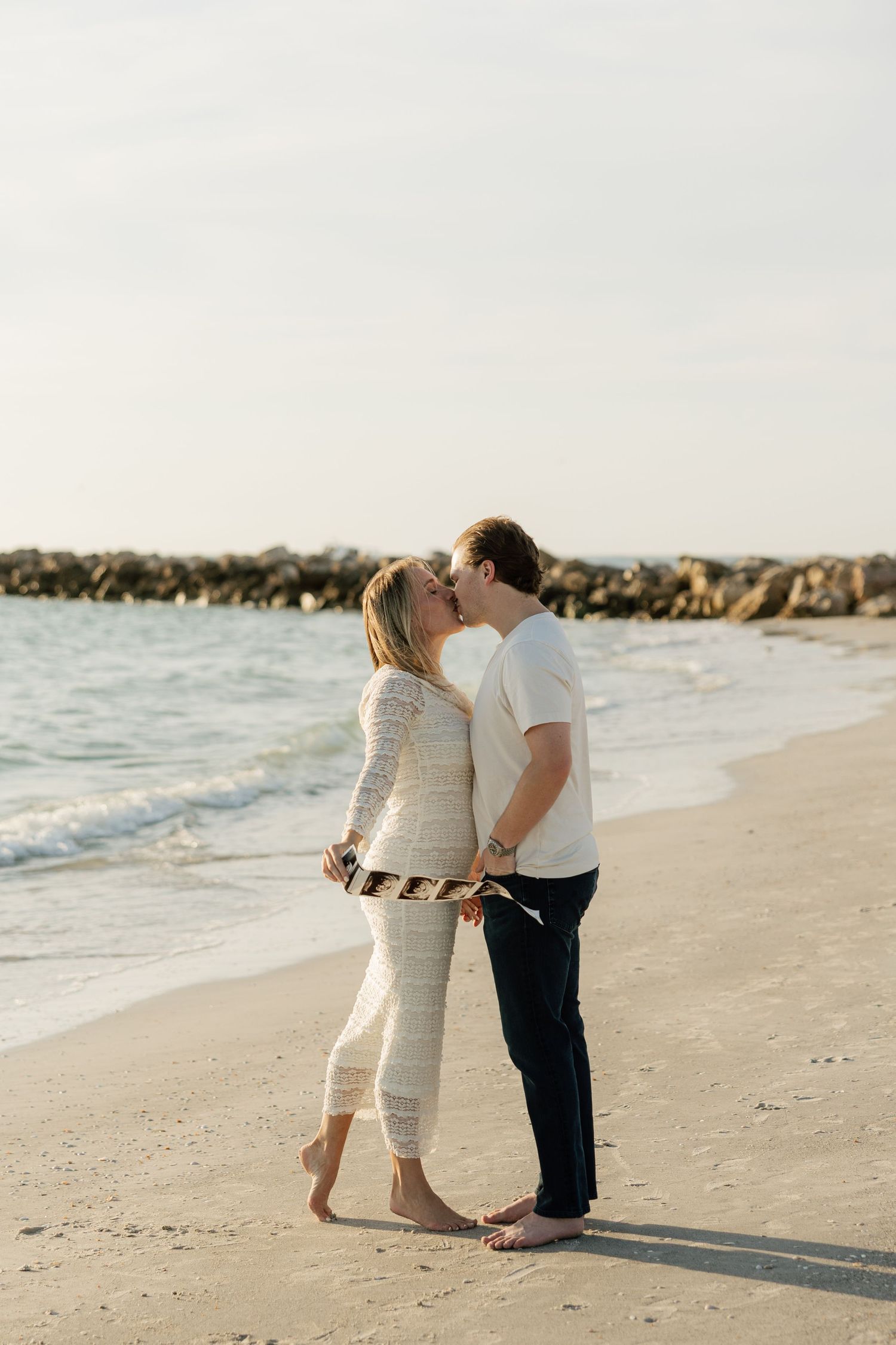 Couple sharing a romantic kiss on a sandy beach with ocean waves and rocky jetty in the background.