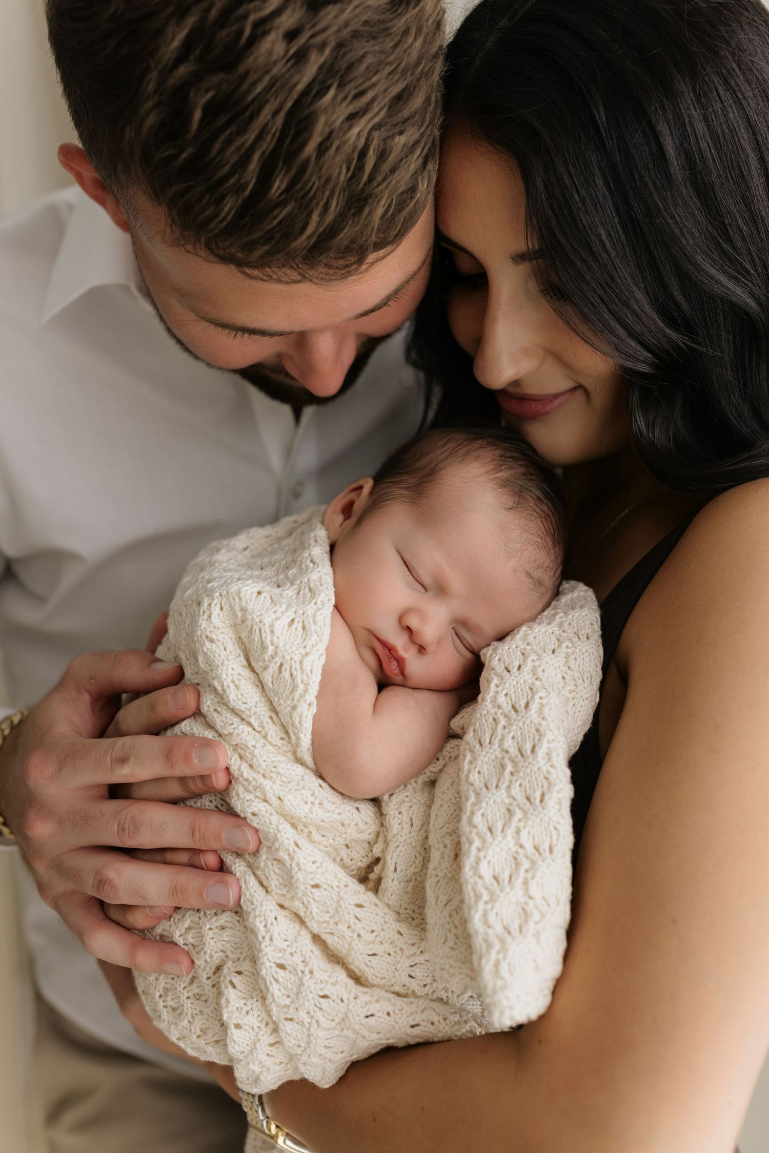 New parents lovingly cradle their sleeping newborn wrapped in a cream knit blanket during a studio portrait session.