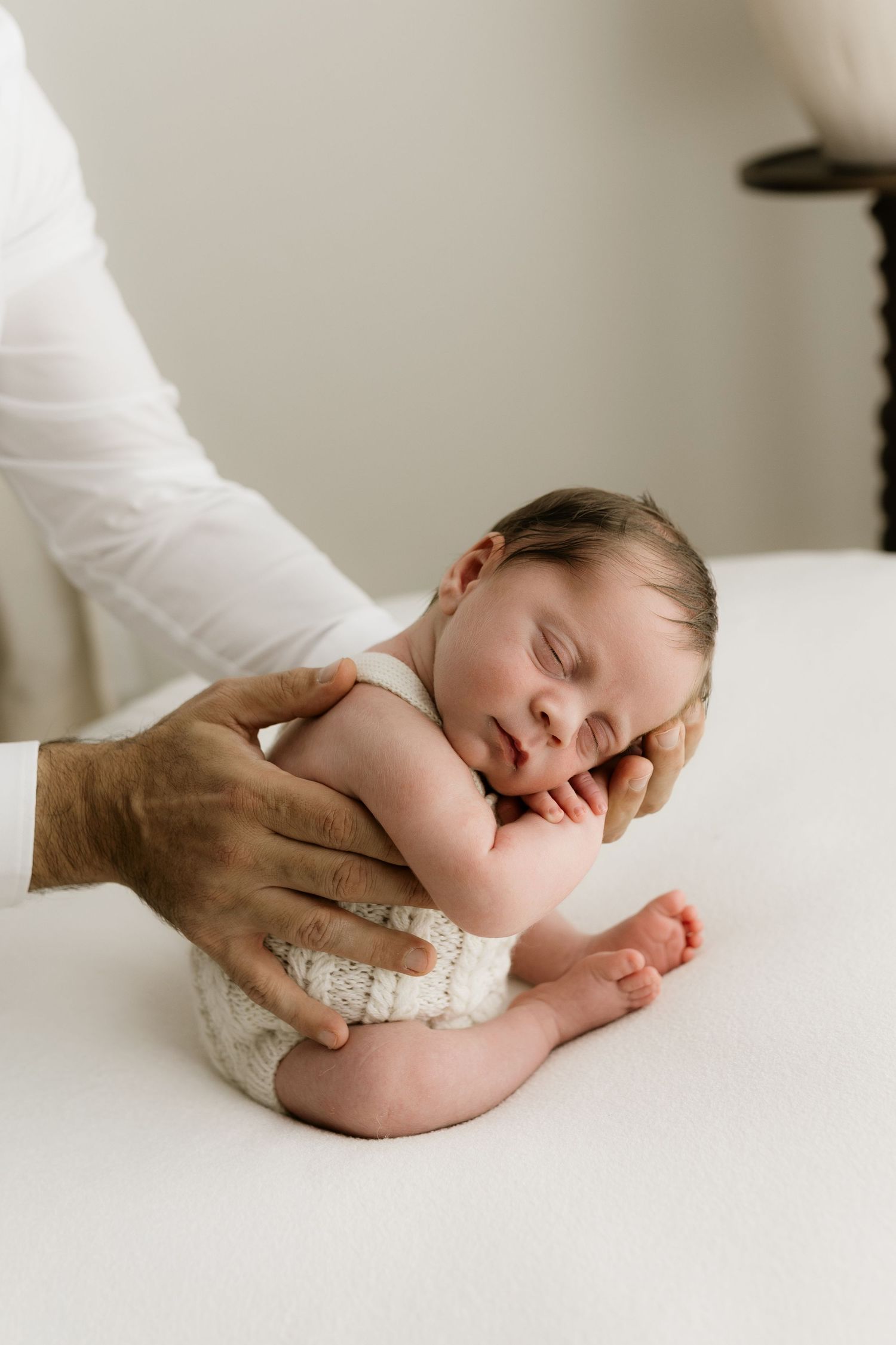 Newborn baby sleeping peacefully while being gently held by adult hands on a white surface.