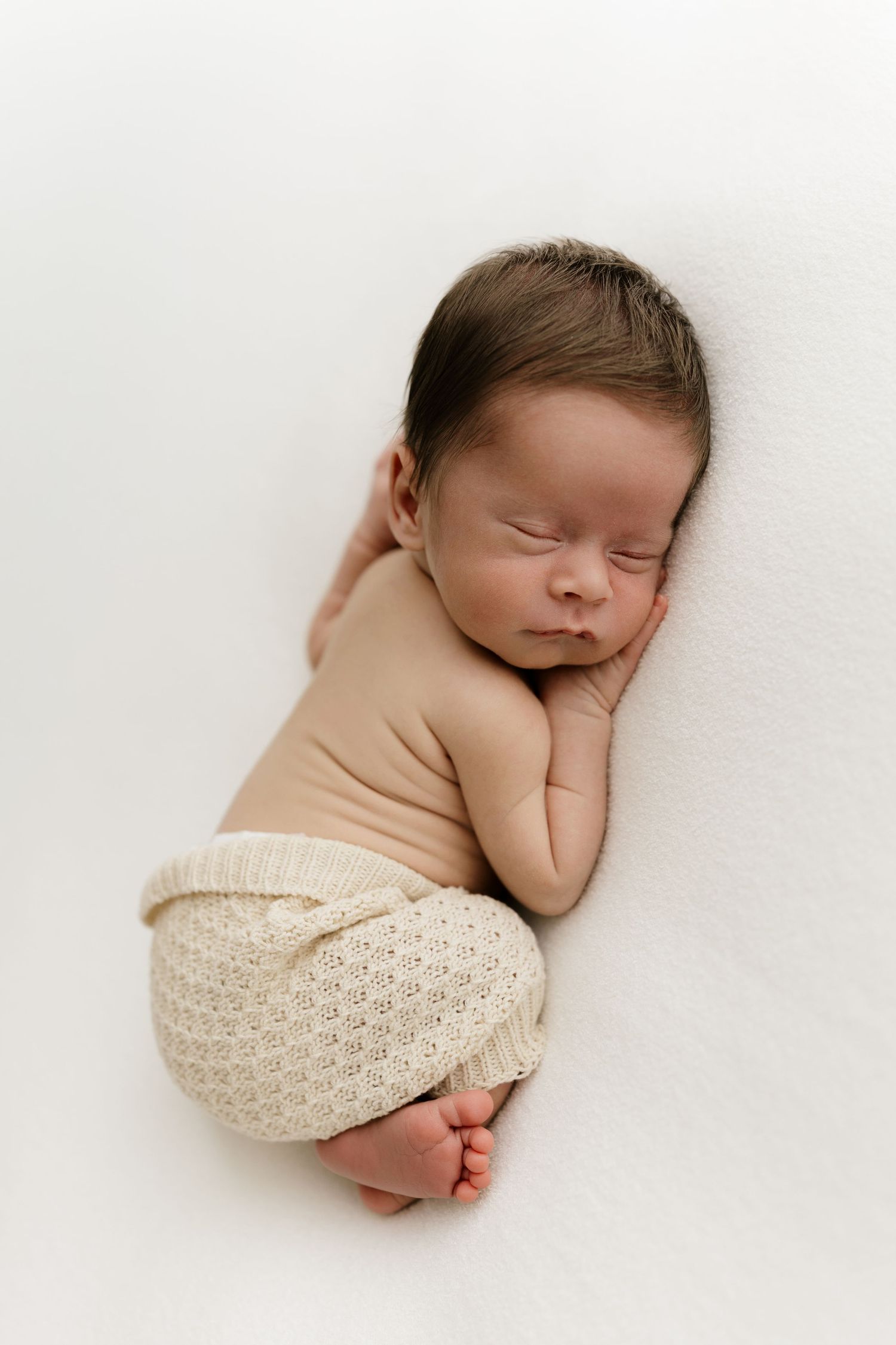 Peaceful newborn baby sleeping curled up on white blanket, wearing beige bodysuit and cream knit shorts.