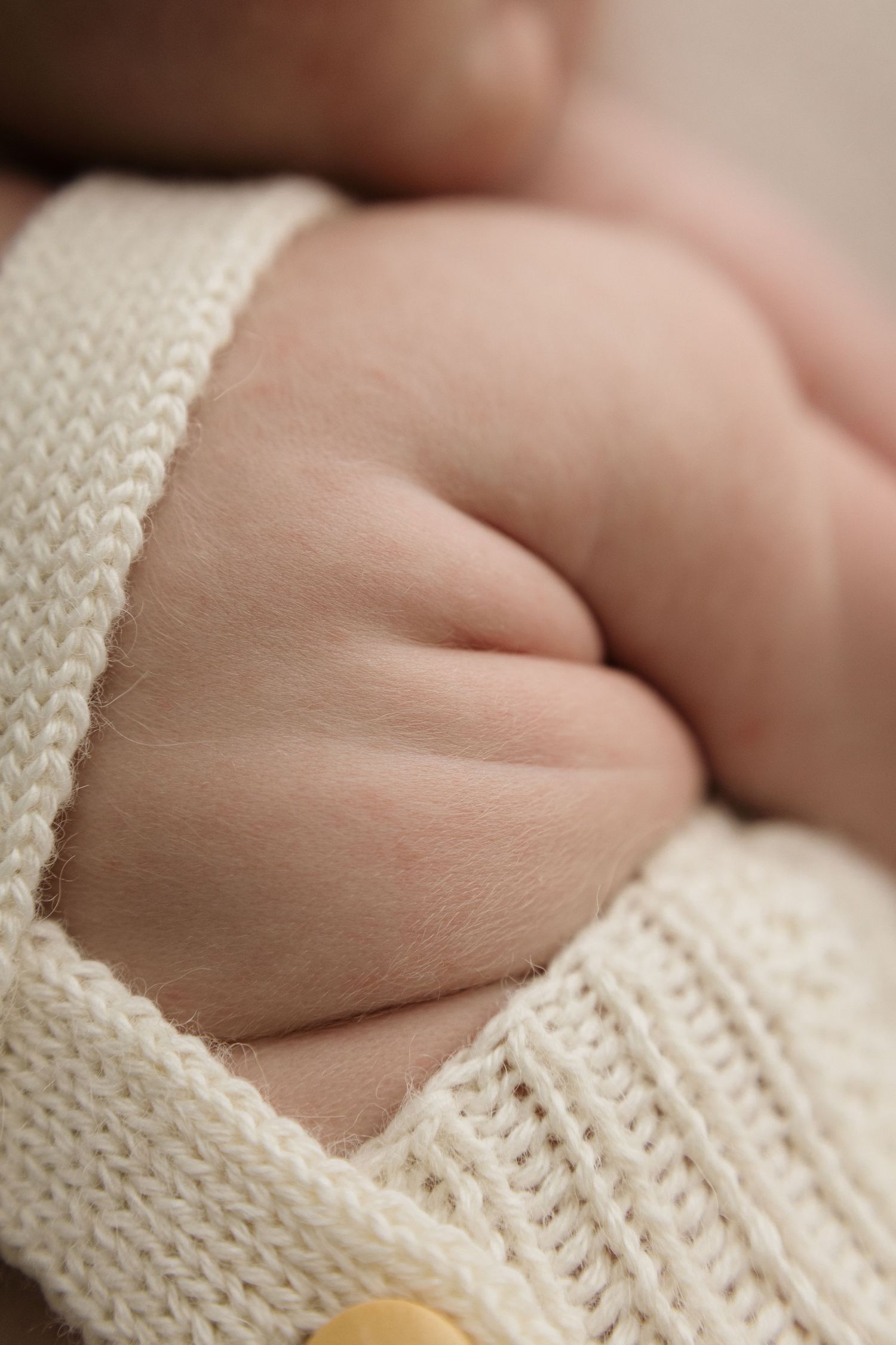 Close-up of a newborn baby's tiny chubby fist resting on a cream knit cardigan sweater.