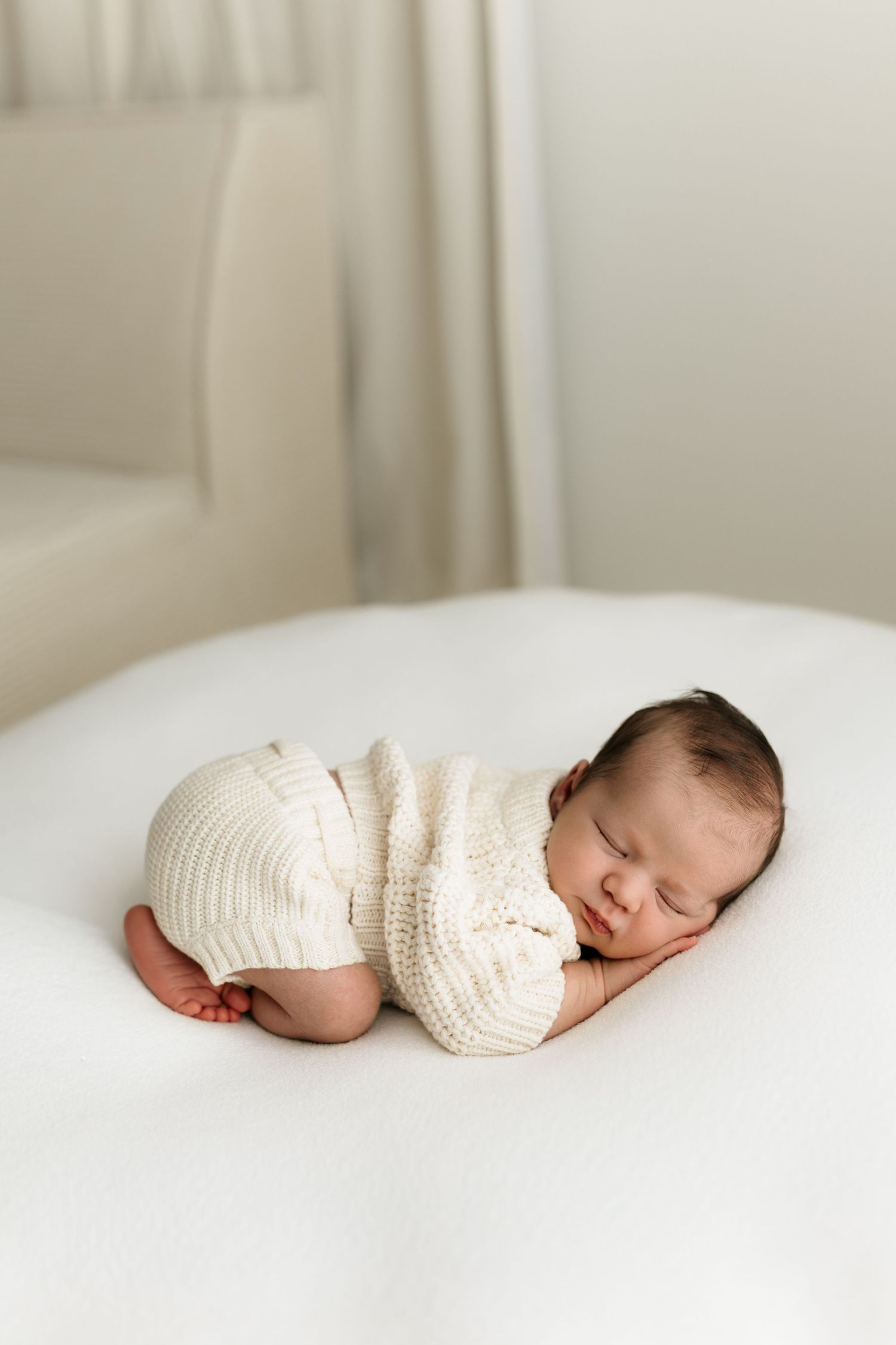 Peaceful newborn baby sleeping curled up in cream knit outfit on white surface.
