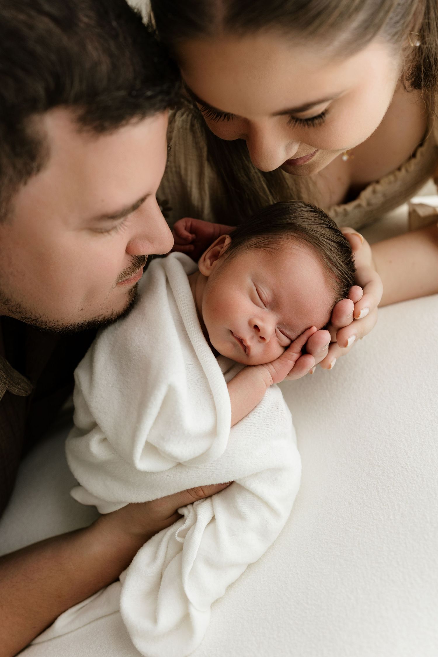 Parents tenderly leaning over their sleeping newborn baby wrapped in a white swaddle blanket.