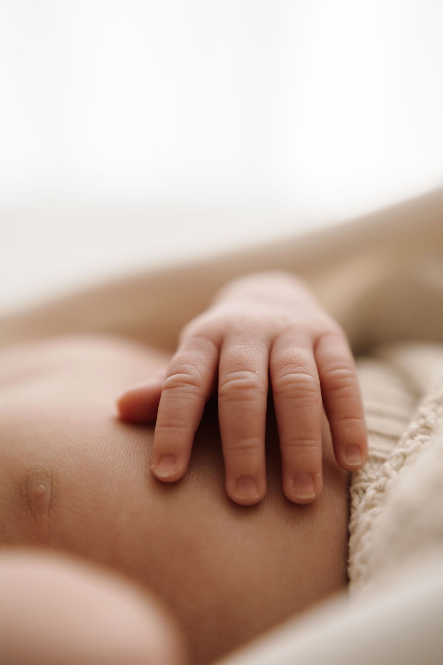 Close-up of a newborn baby's tiny delicate hand resting gently on their stomach, wrapped in a soft beige knit blanket.