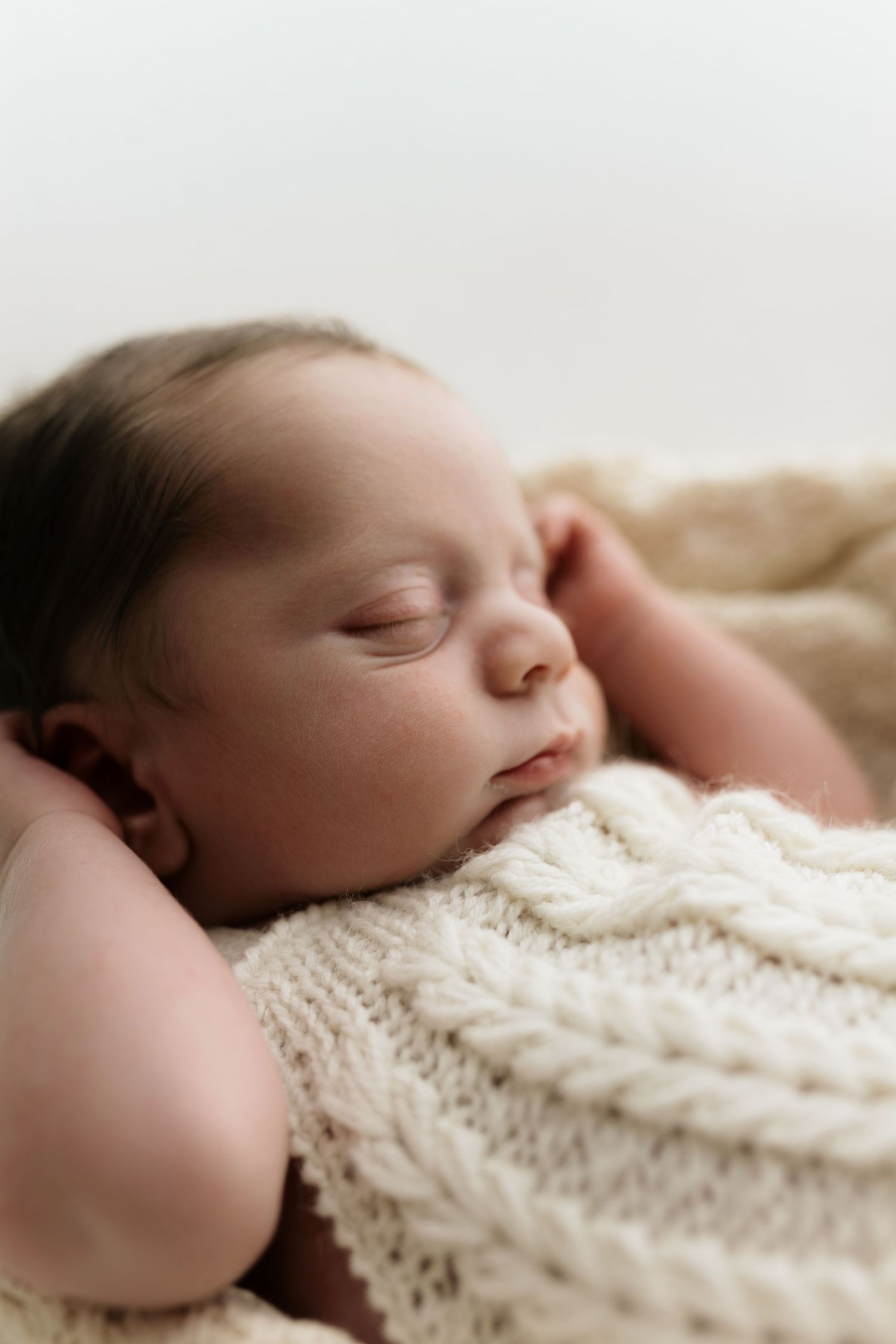 Peaceful newborn baby sleeping soundly on a soft cream knit blanket with tiny hands raised beside face.