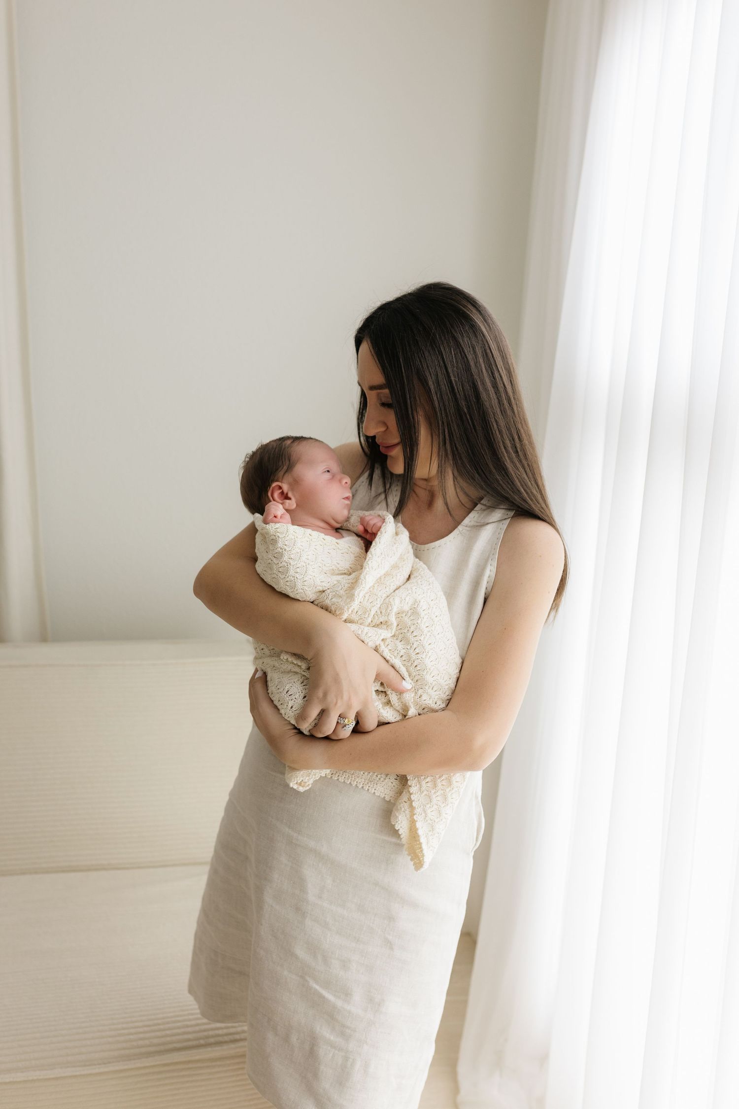 A mother in a white linen dress tenderly holds her swaddled newborn baby near a bright window.