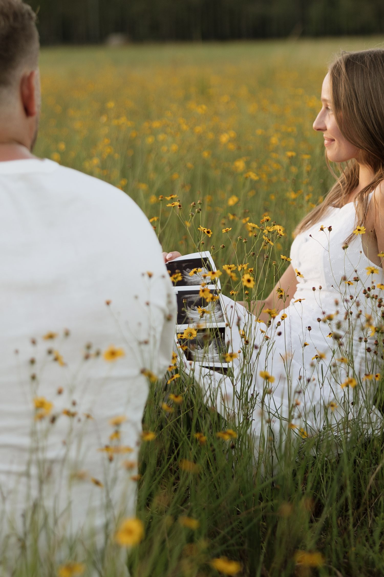 Pregnant woman in white dress sitting in yellow wildflower field, sharing ultrasound photos with partner.
