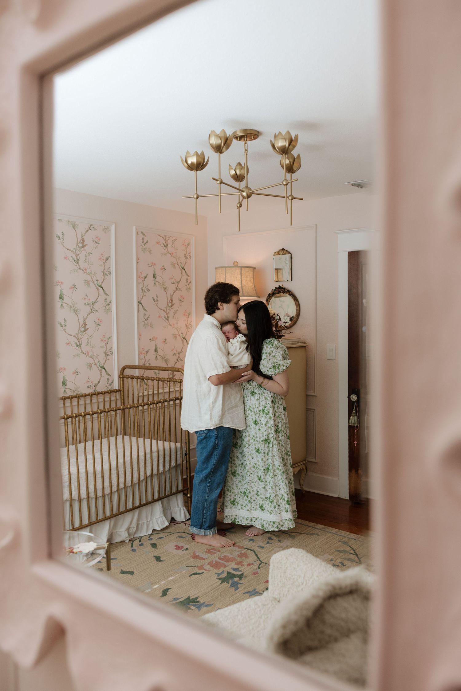 A couple shares a tender moment with their newborn in a soft pink nursery with floral wallpaper, gold crib, and chandelier.