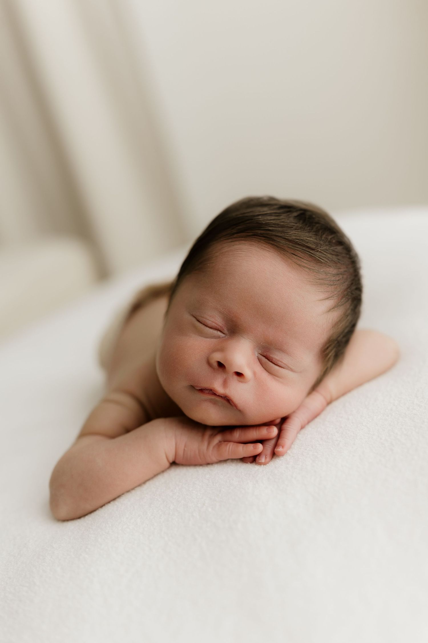Peaceful newborn baby sleeping with hands tucked under chin on a soft white blanket in a studio portrait.