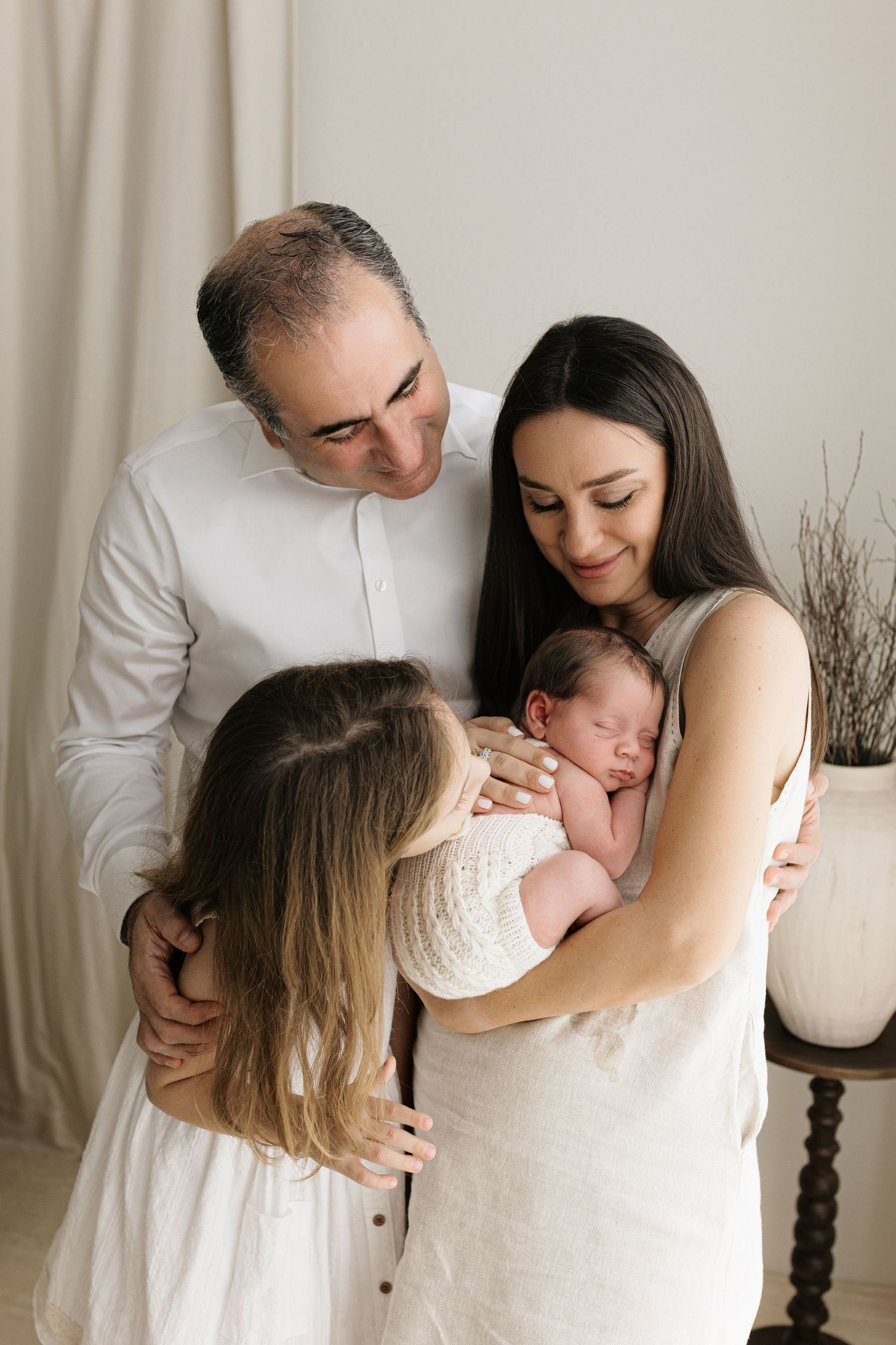 A family of four gathers lovingly around their newborn baby, with parents and young daughter sharing a tender moment together.