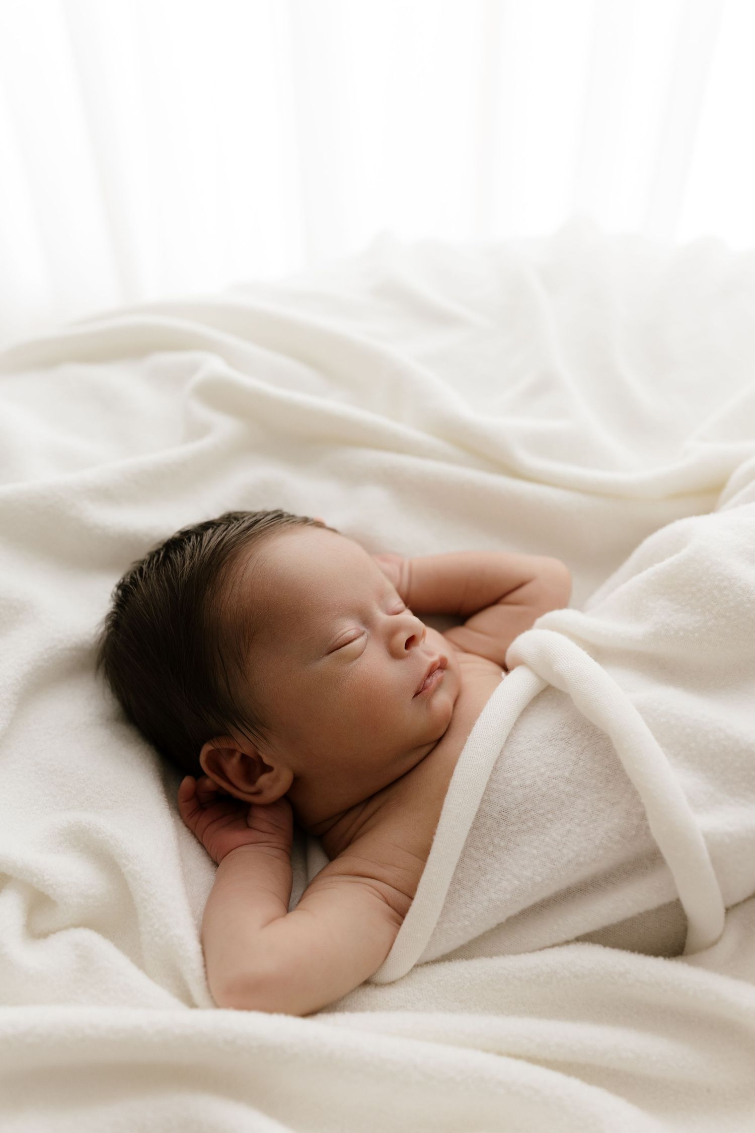 Serene newborn baby sleeping wrapped in a soft white blanket with natural window light in background.