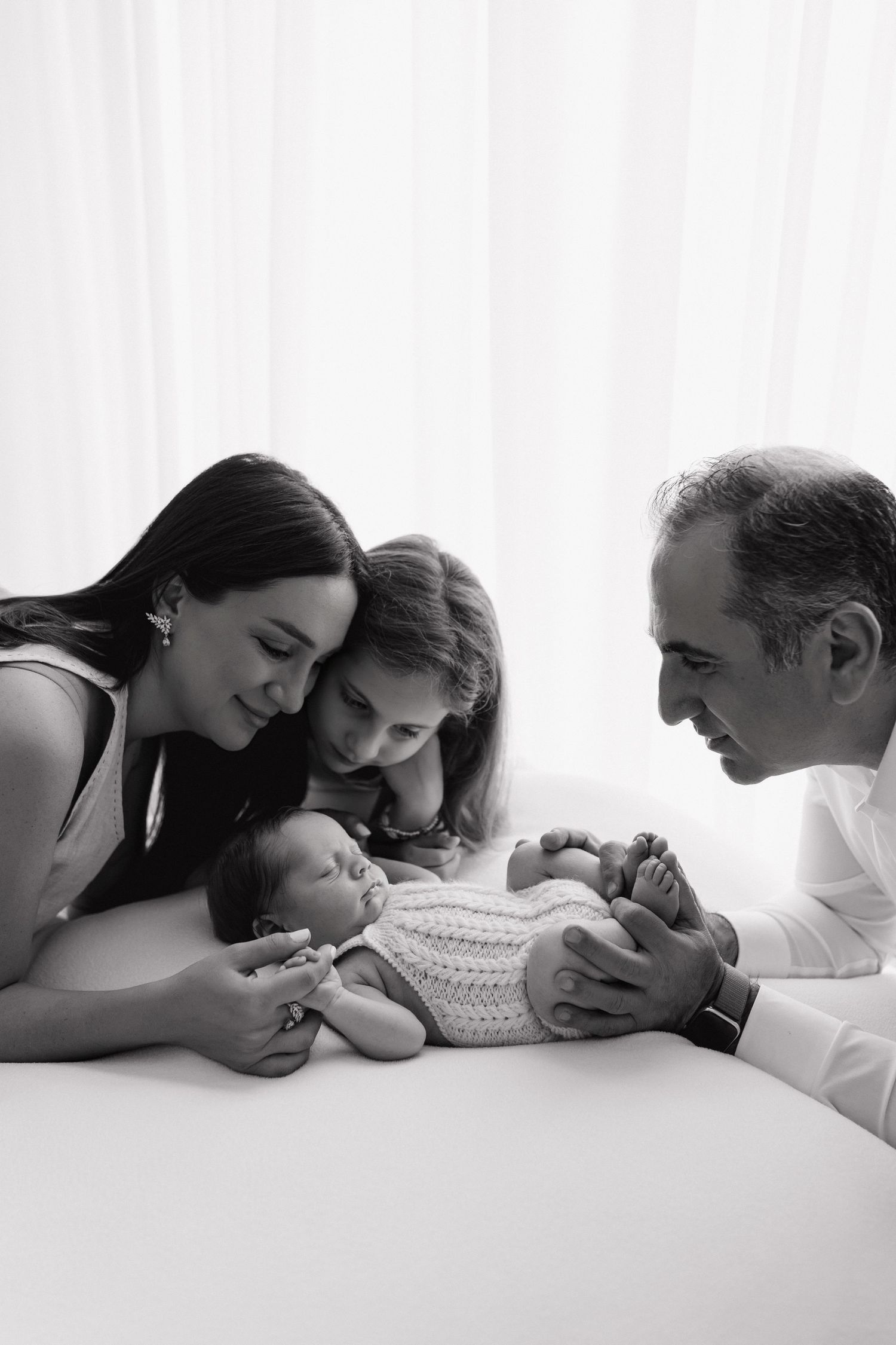 Black and white family photo of parents and sibling lovingly gazing at sleeping newborn baby on white surface.
