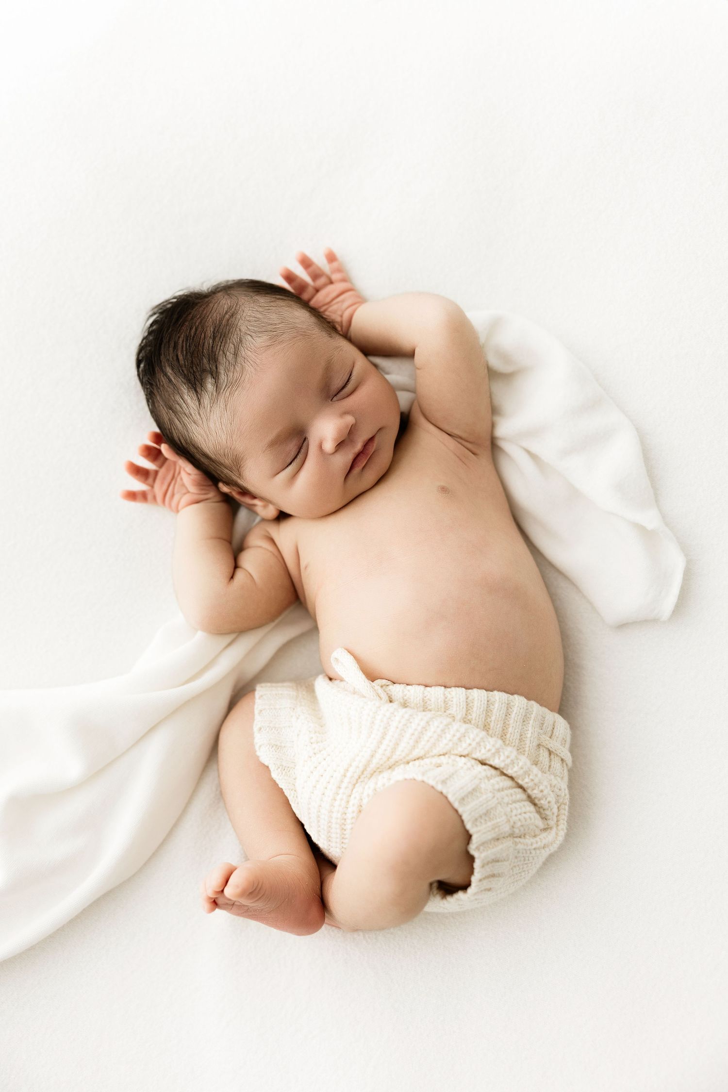 Peaceful newborn baby sleeping on white blanket wearing cream ribbed shorts.