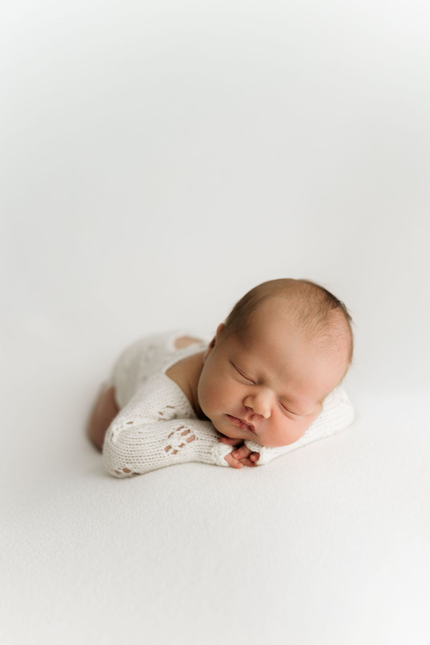 Peaceful newborn baby sleeping in white knit romper on soft white blanket in professional studio portrait.
