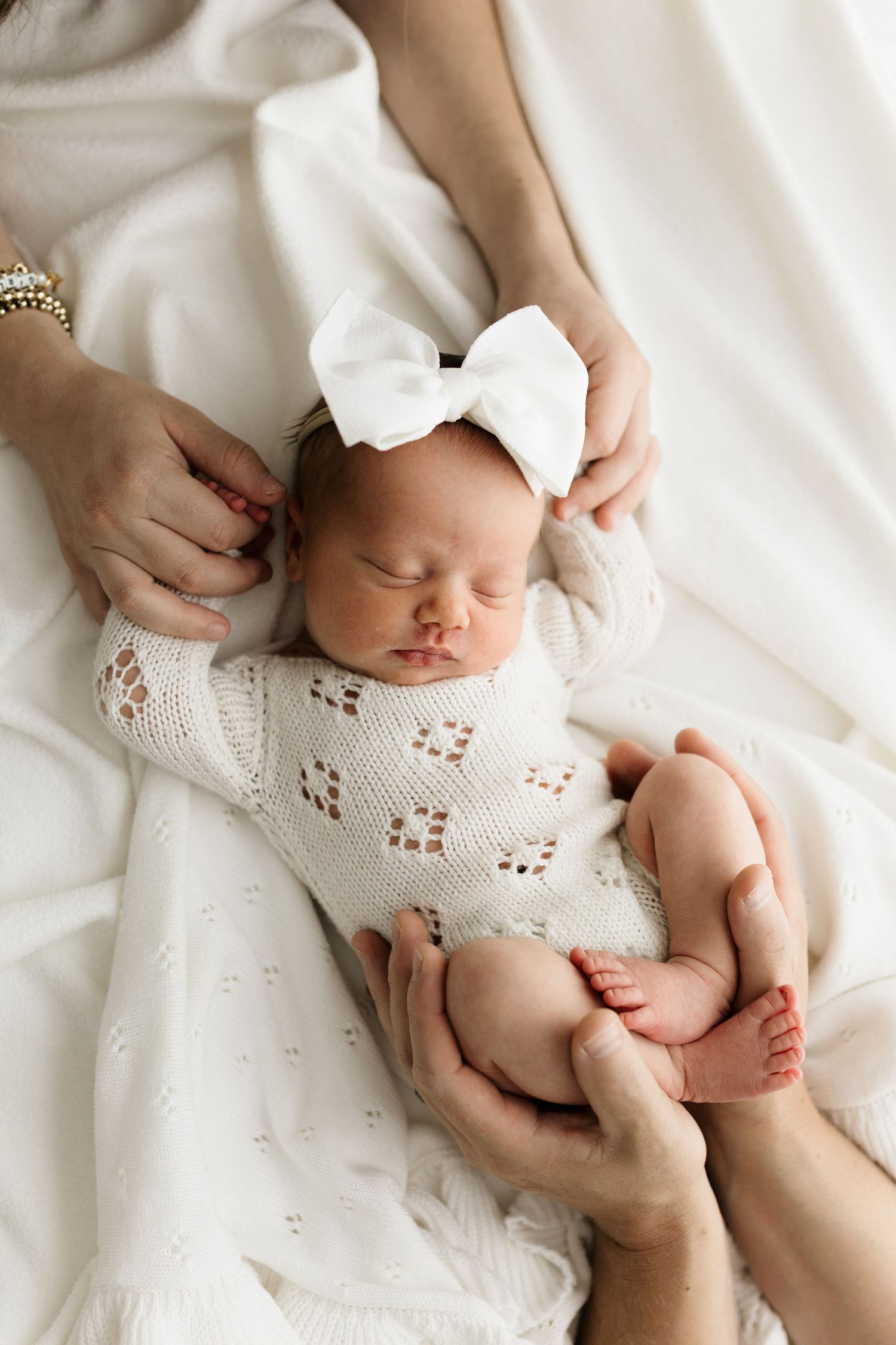 Parents gently holding their sleeping newborn baby girl in a white knit outfit with a bow headband.