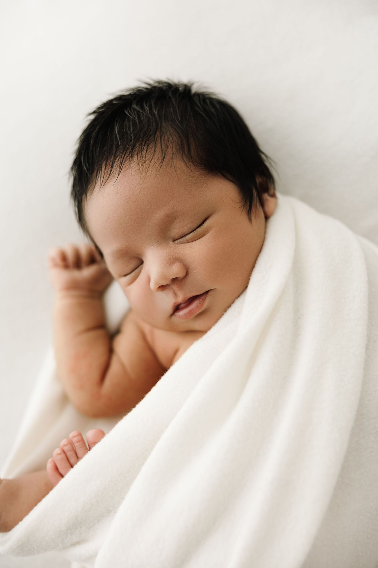 A peaceful sleeping newborn baby swaddled in soft white blankets rests against a light background.