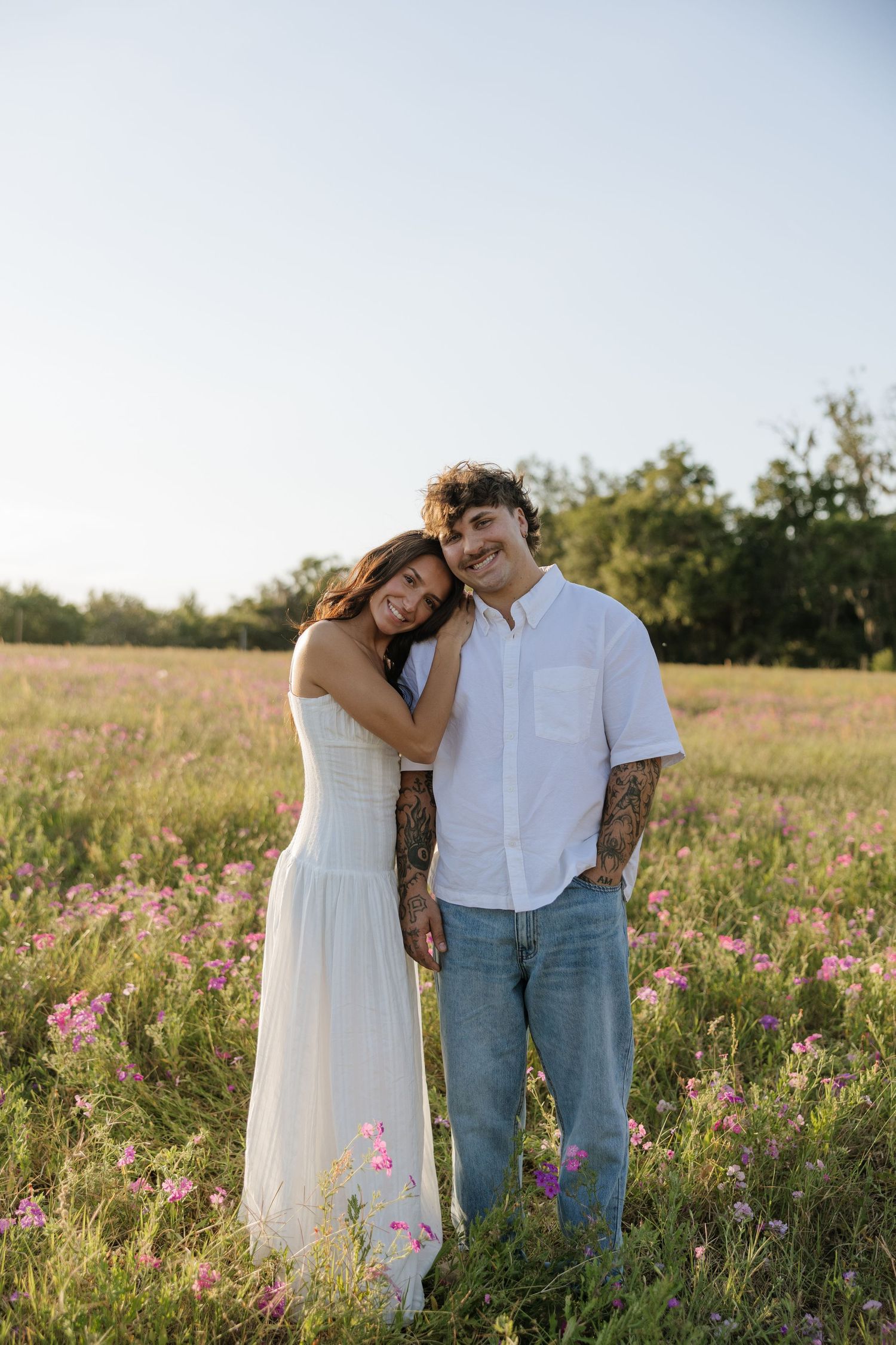 Couple embracing in a wildflower field, woman in white dress leaning on tattooed man in white shirt and jeans.