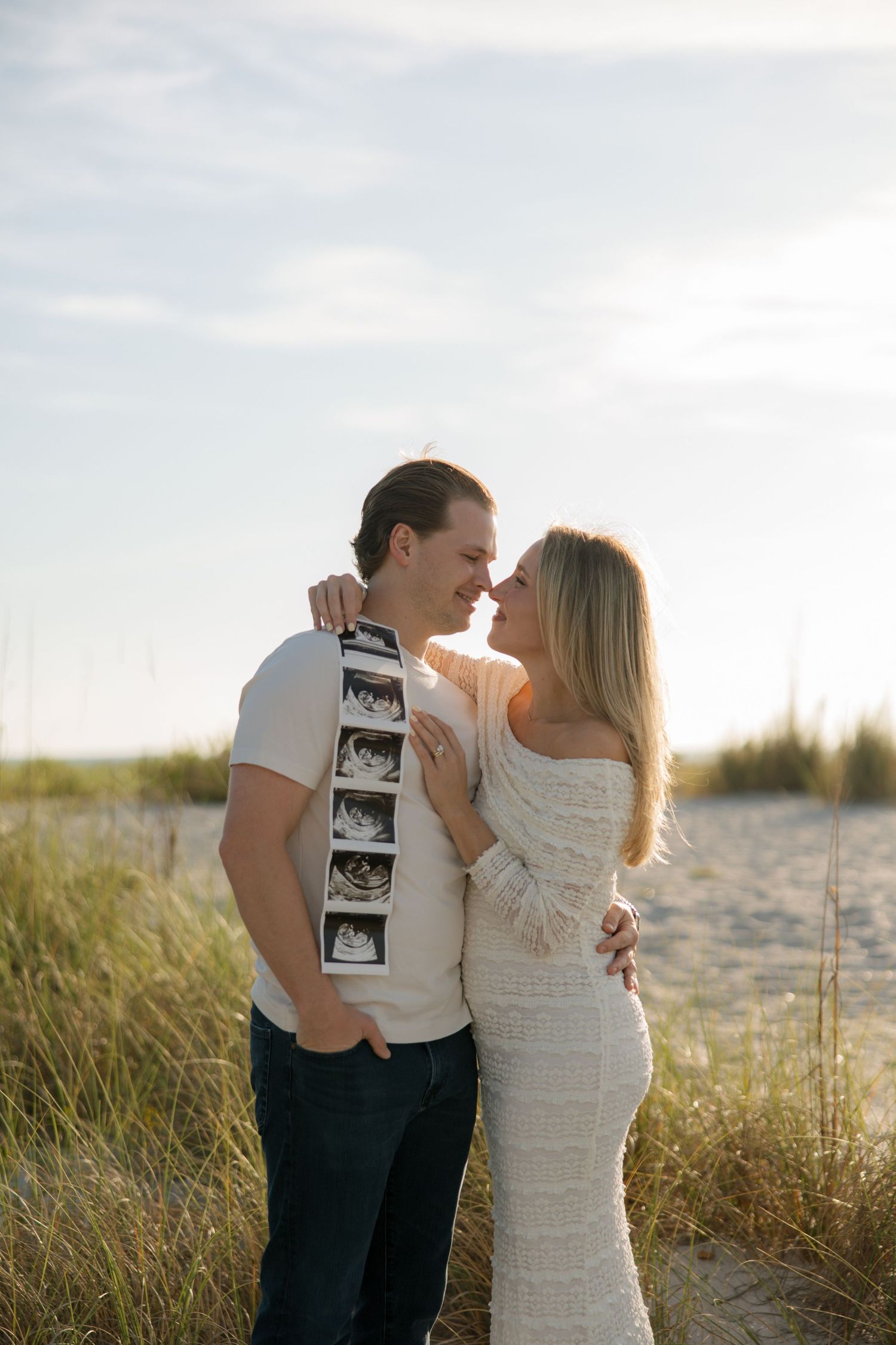 Couple embracing on beach holding ultrasound photos, announcing pregnancy at sunset among sand dunes.