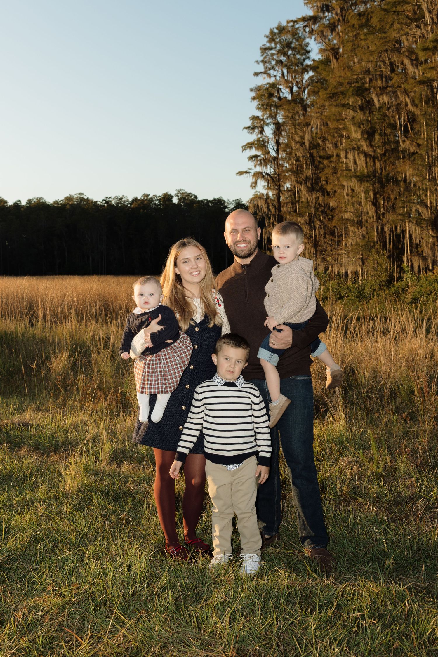 Family group playing together in grassy field at sunset near wooded area.