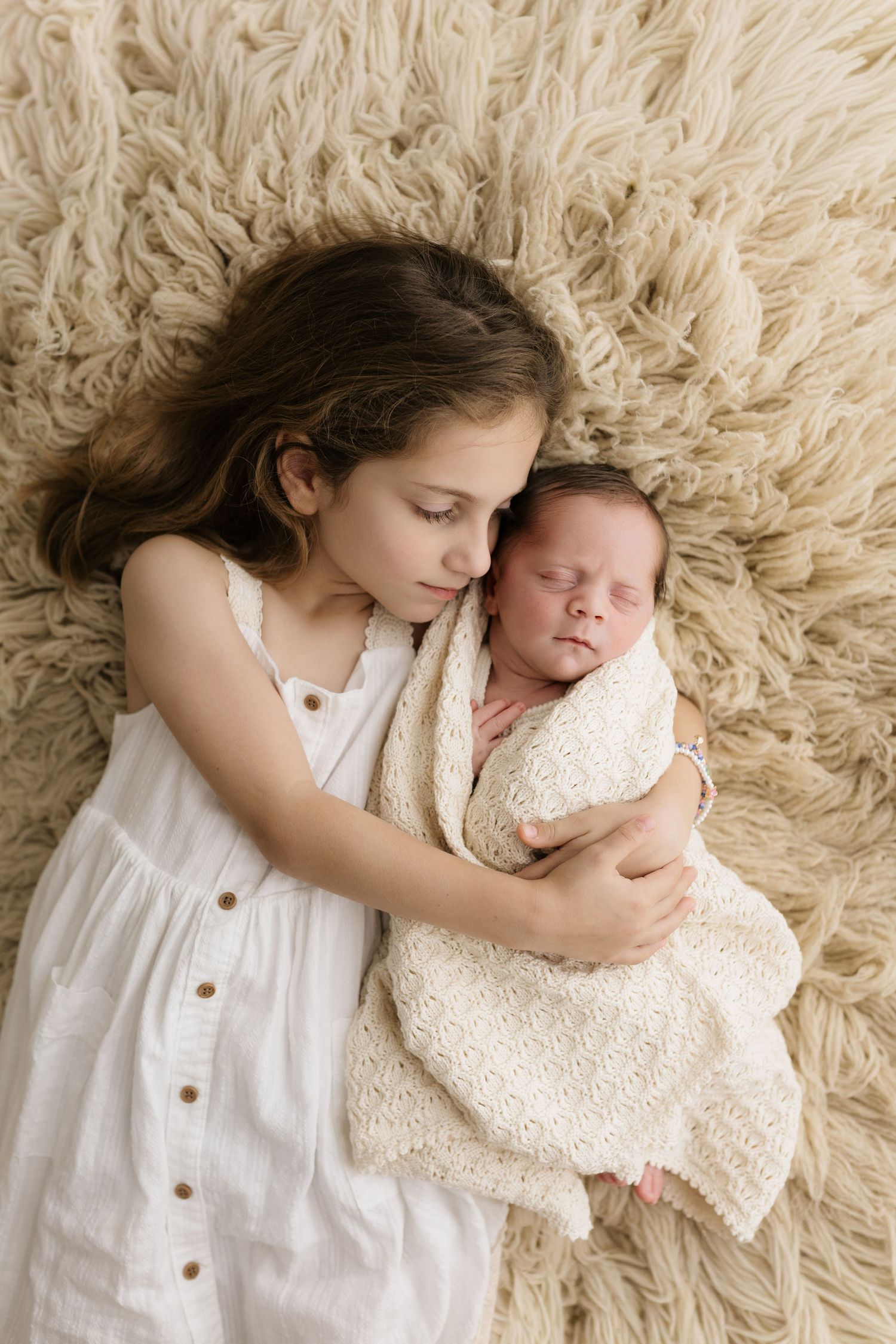 Young girl in white dress tenderly holds sleeping newborn sibling on fluffy beige rug in sweet portrait.