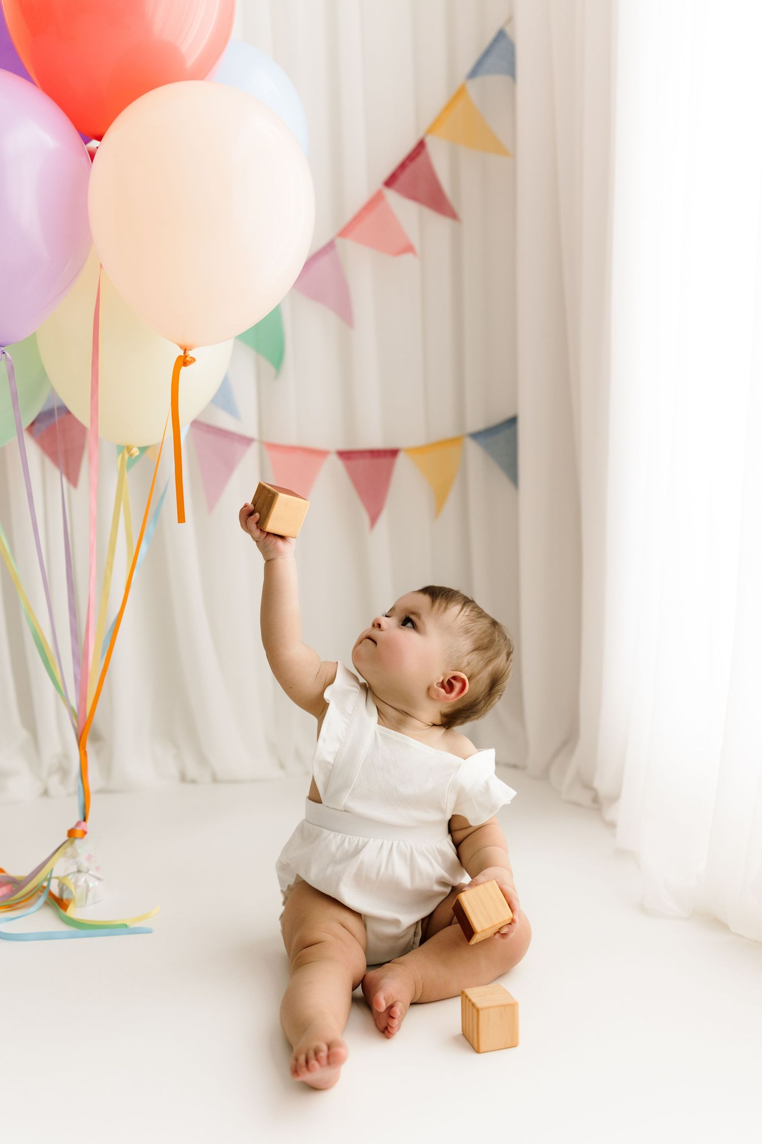 A baby in a white outfit sits on the floor holding wooden blocks near colorful balloons and party bunting decorations.
