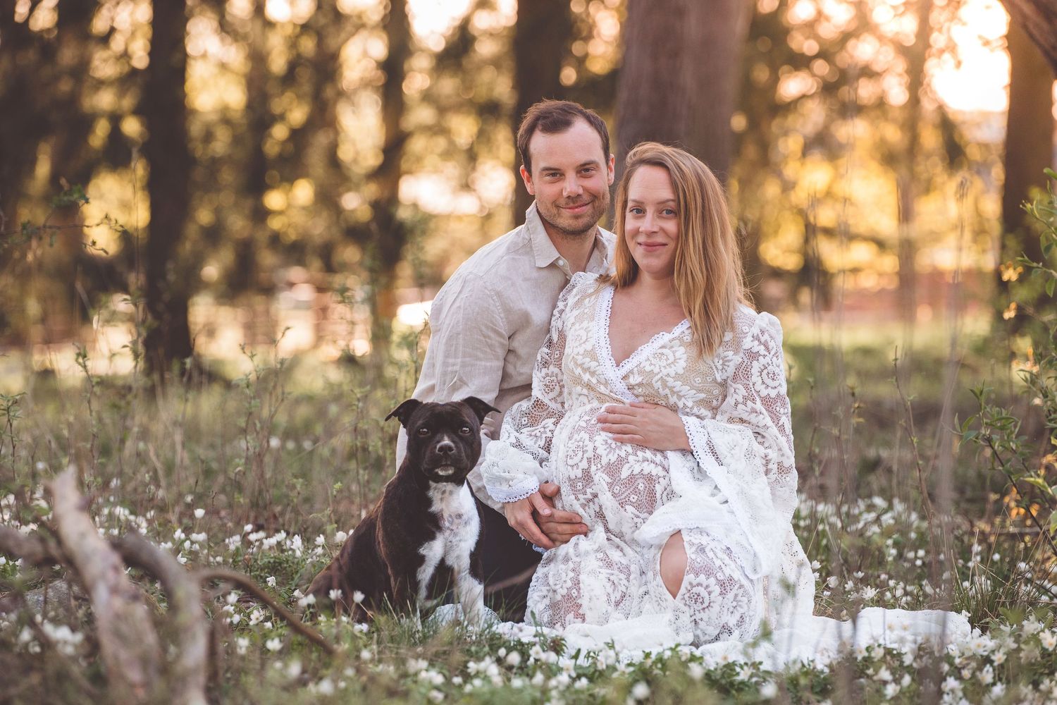 Couple in wedding attire embraces in a wildflower meadow at sunset while their black dog watches nearby.