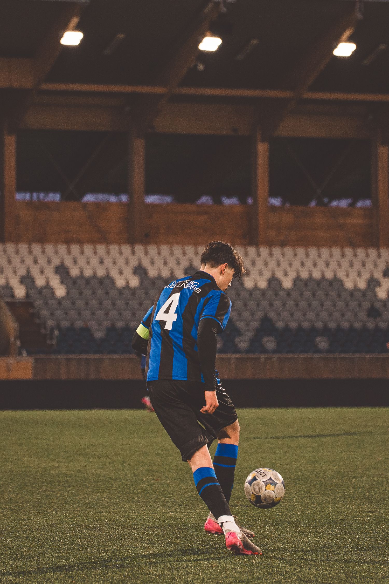 Soccer player in blue and black stripes dribbles ball across empty stadium field.