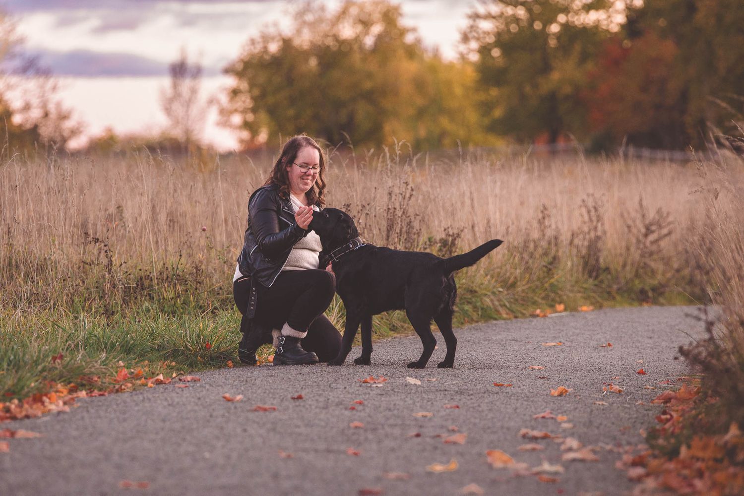 A black dog and their companion walk together on a path during autumn as fallen leaves scatter around them.