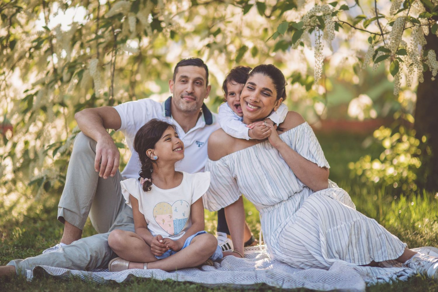 Family enjoying a sunny afternoon picnic together on a white blanket under blooming trees in a park.