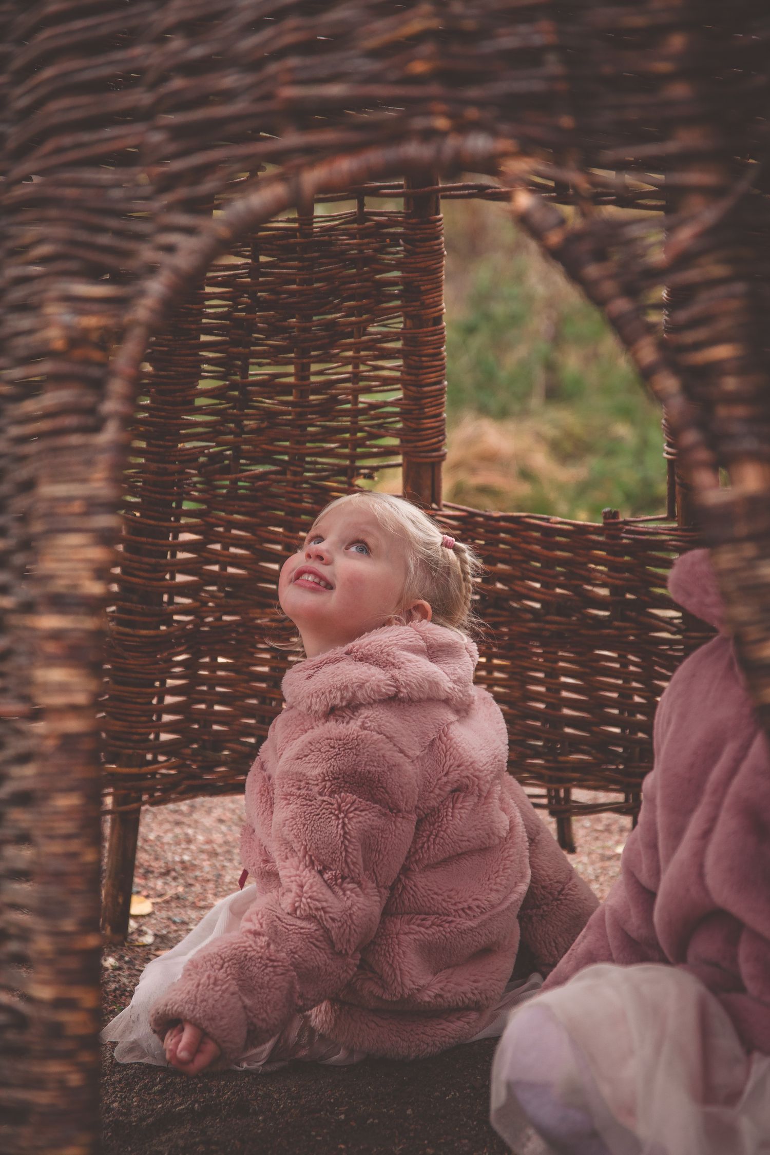Young child in a pink fuzzy coat plays in a woven willow structure during autumn.