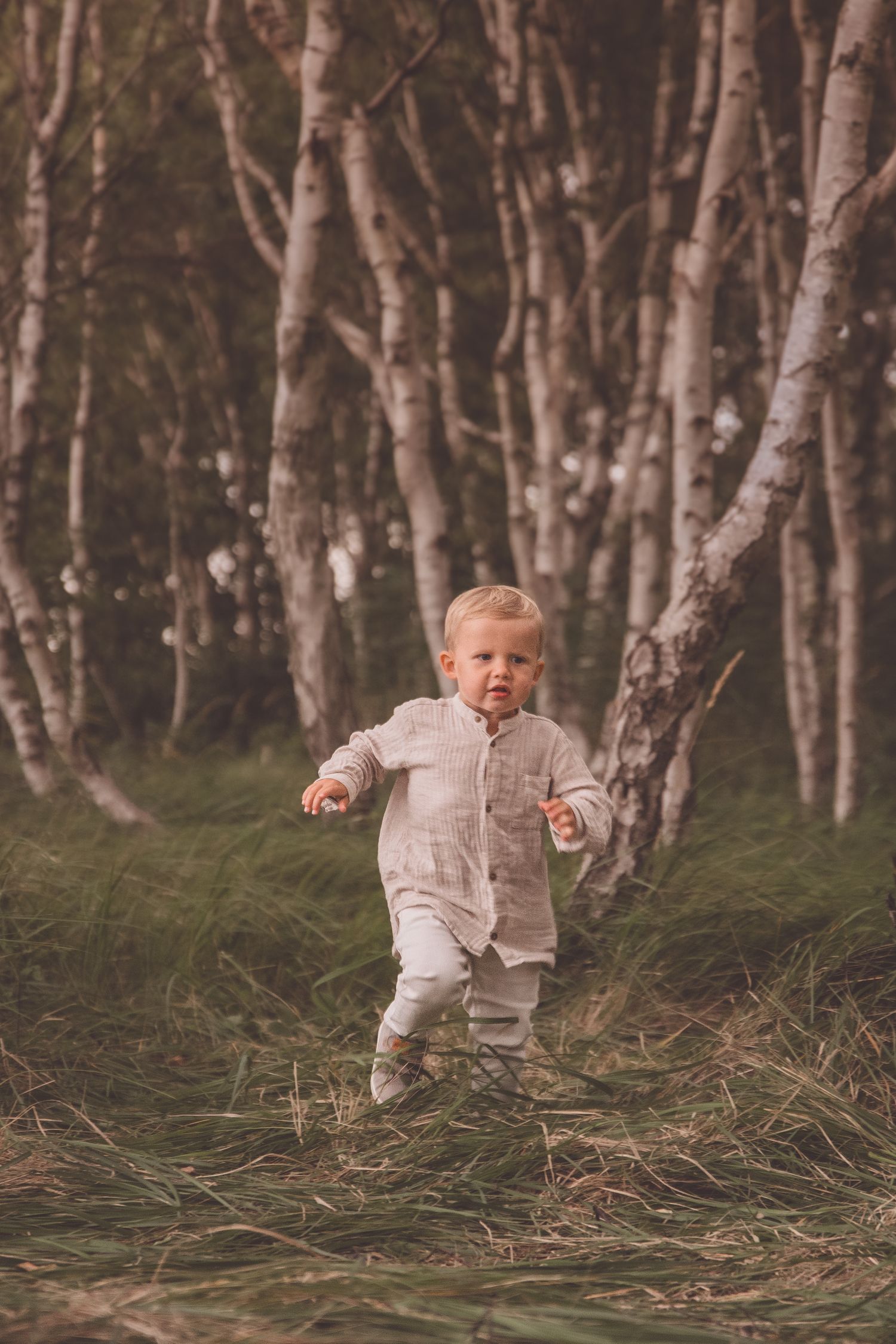 A child in white clothing runs joyfully through a birch forest with tall trees and grassy ground.