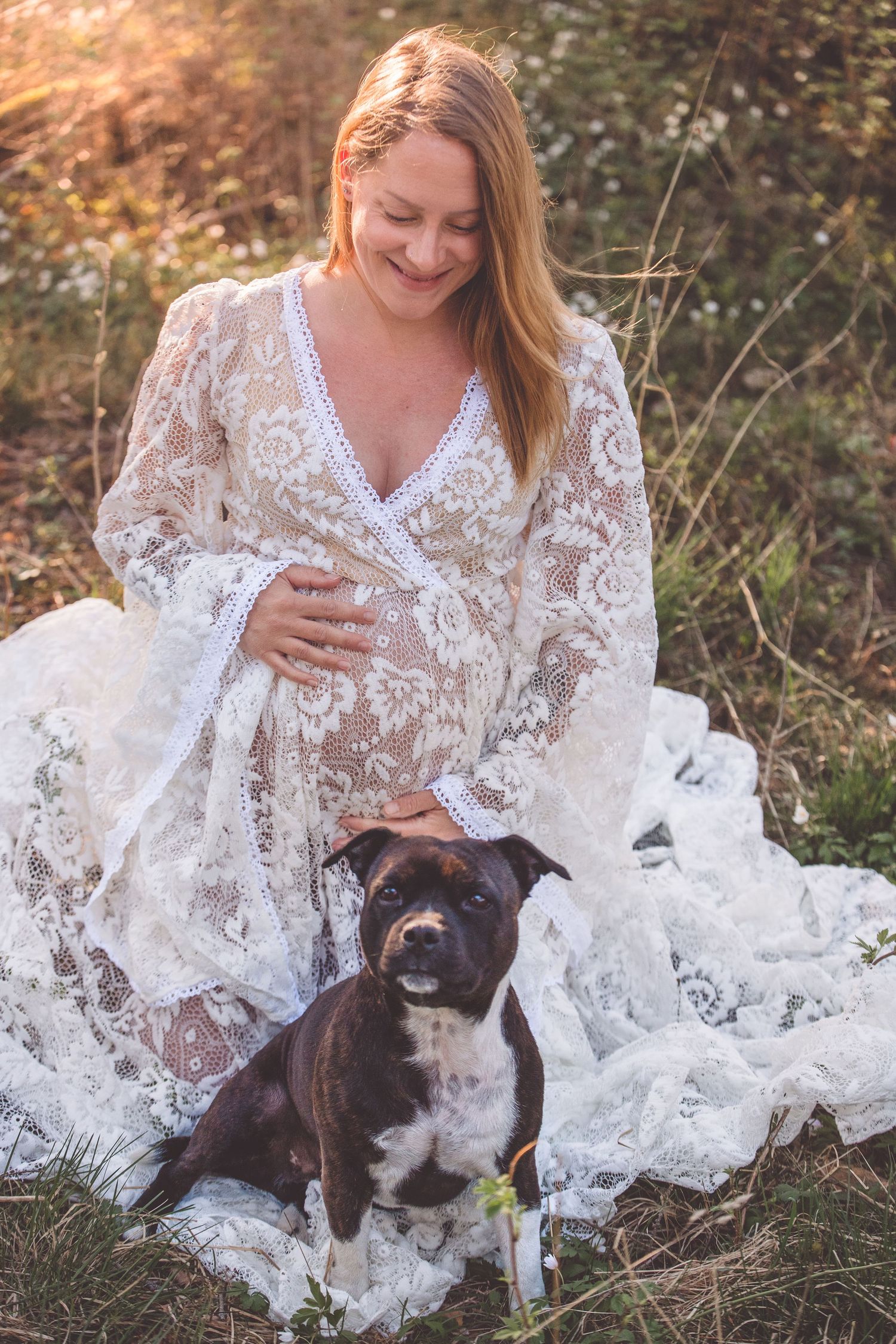 A person in a white lace dress sits in a field with their black and white dog during a golden sunset.