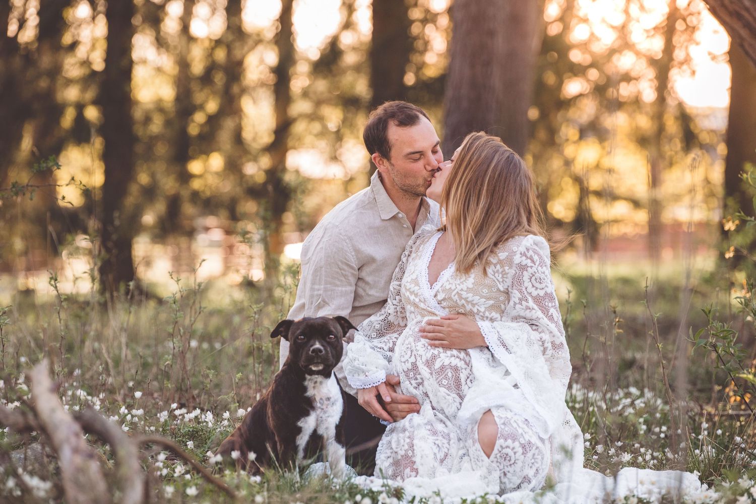 Couple embraces in a wildflower meadow at golden sunset with their dog beside them.