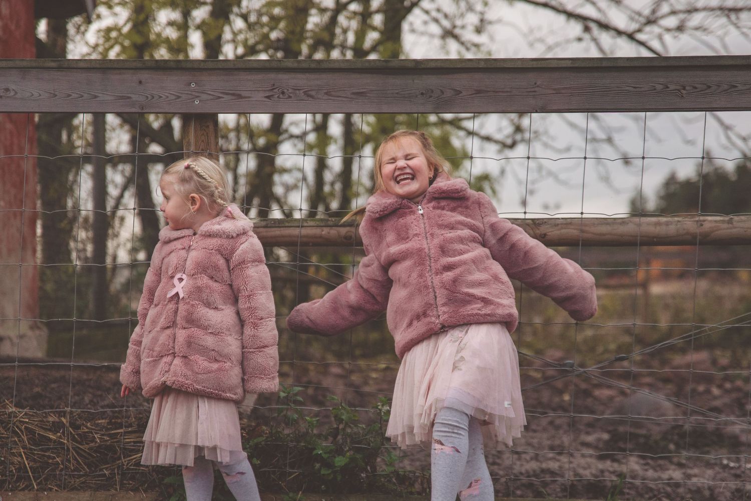 Children in matching pink jackets play together near a wooden fence during autumn.