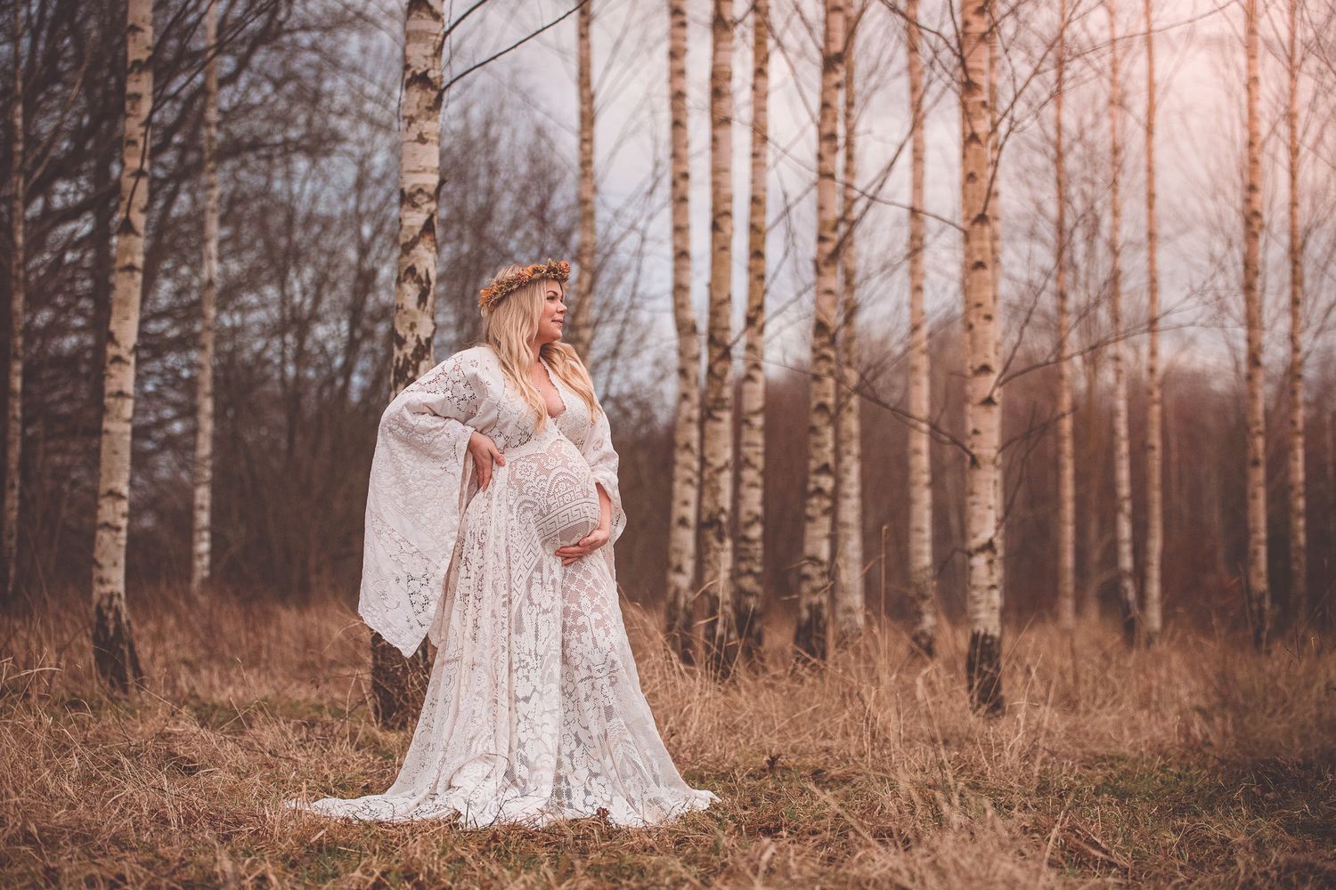 A dreamy maternity portrait in a white lace dress among birch trees at sunset in an autumn forest.