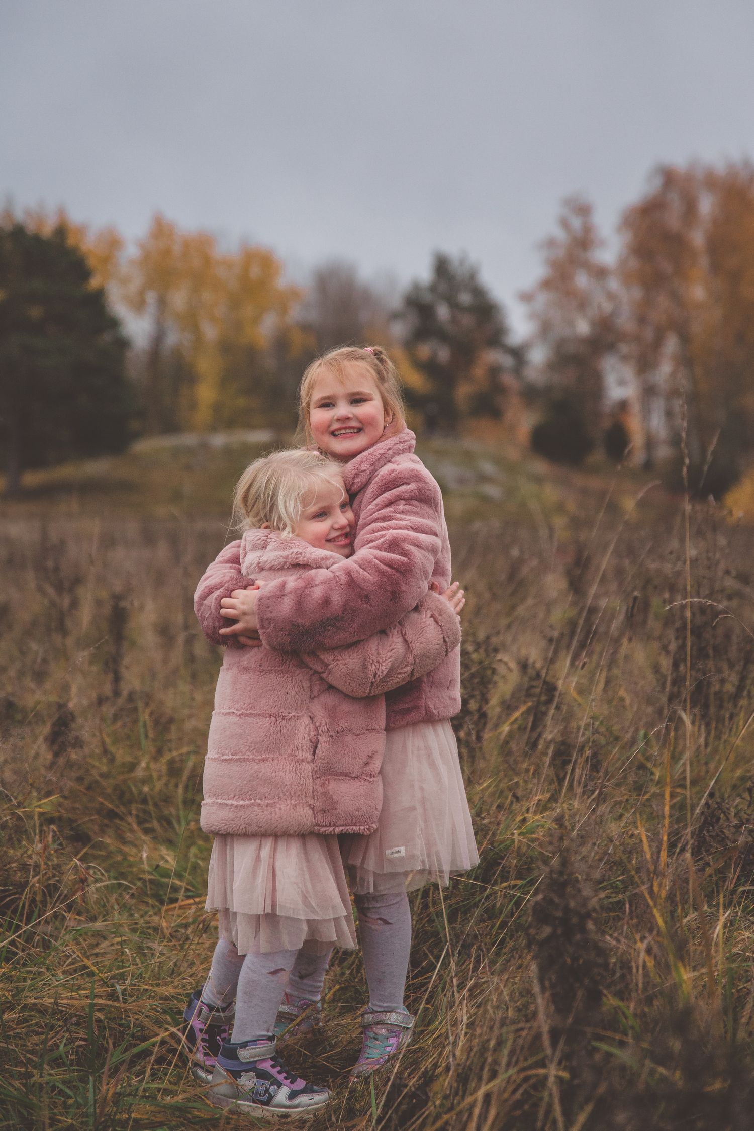 Two children in pink coats embrace while standing in tall autumn grass.