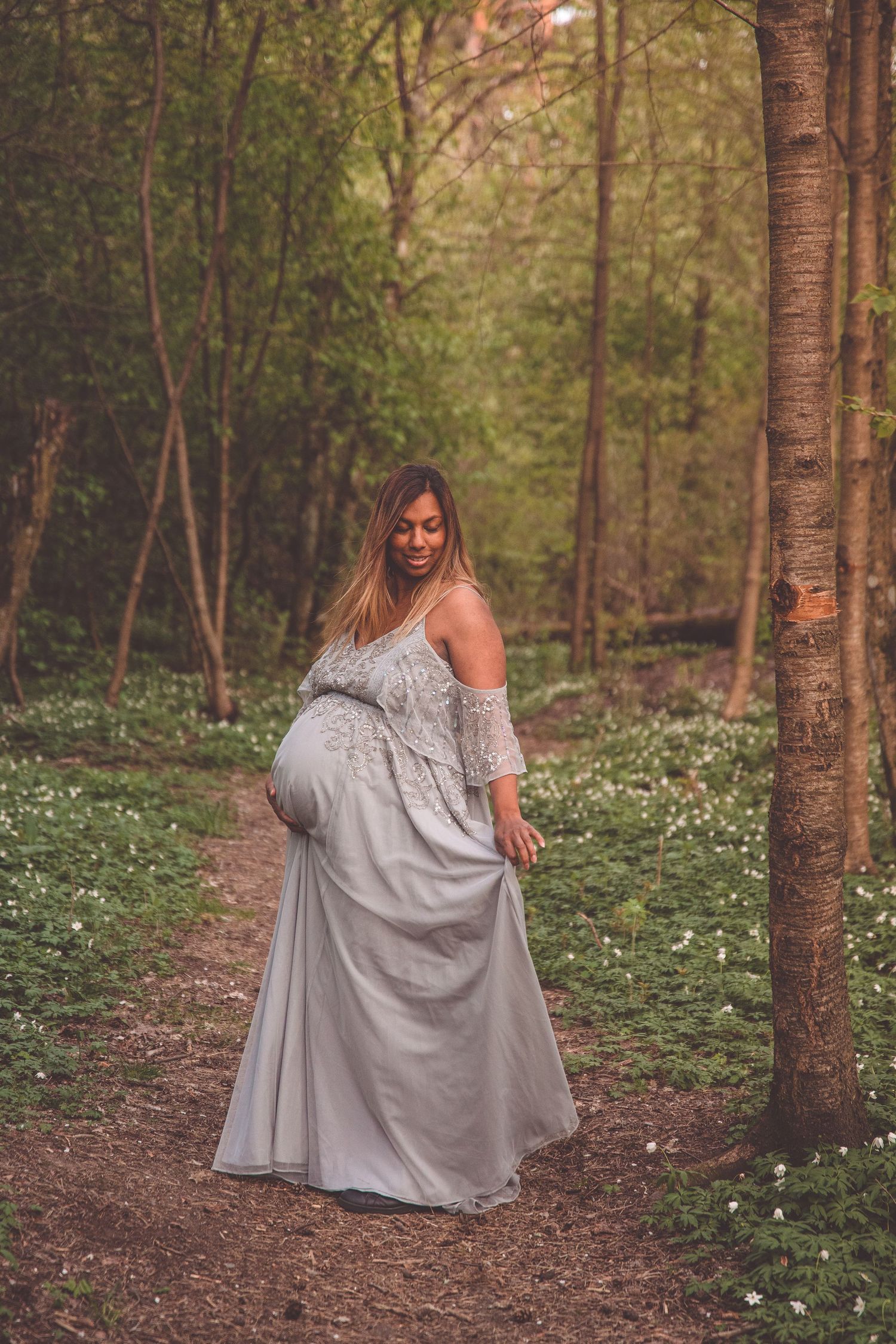 A flowing white off-shoulder maternity dress worn in a serene forest setting with white wildflowers dotting the ground.