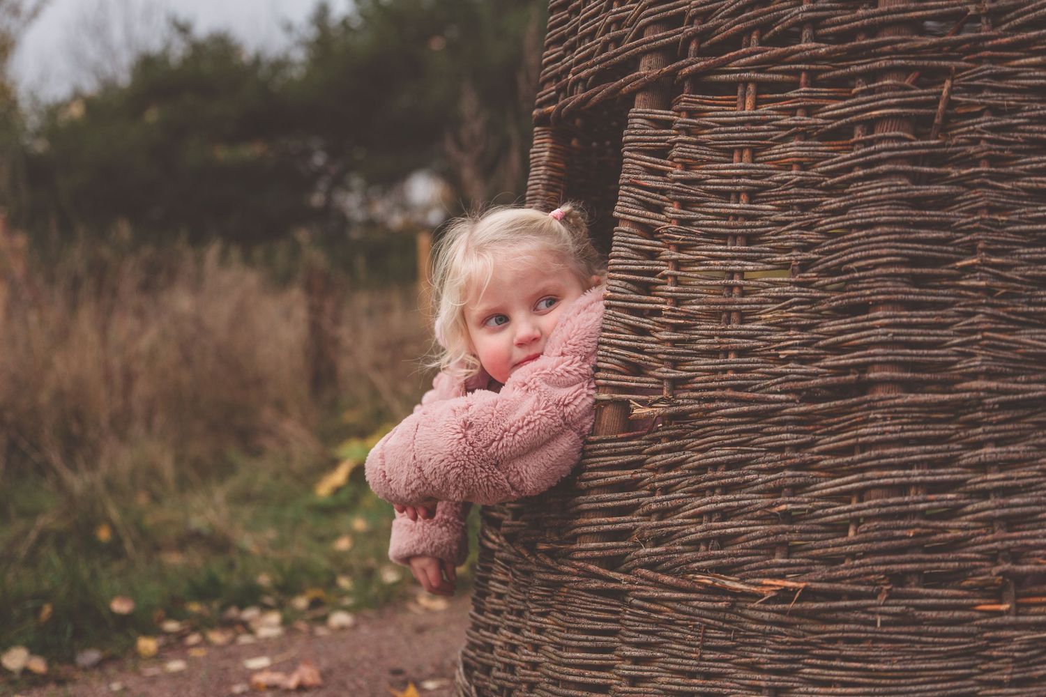 Children in pink coats peek around a woven wicker structure outdoors.