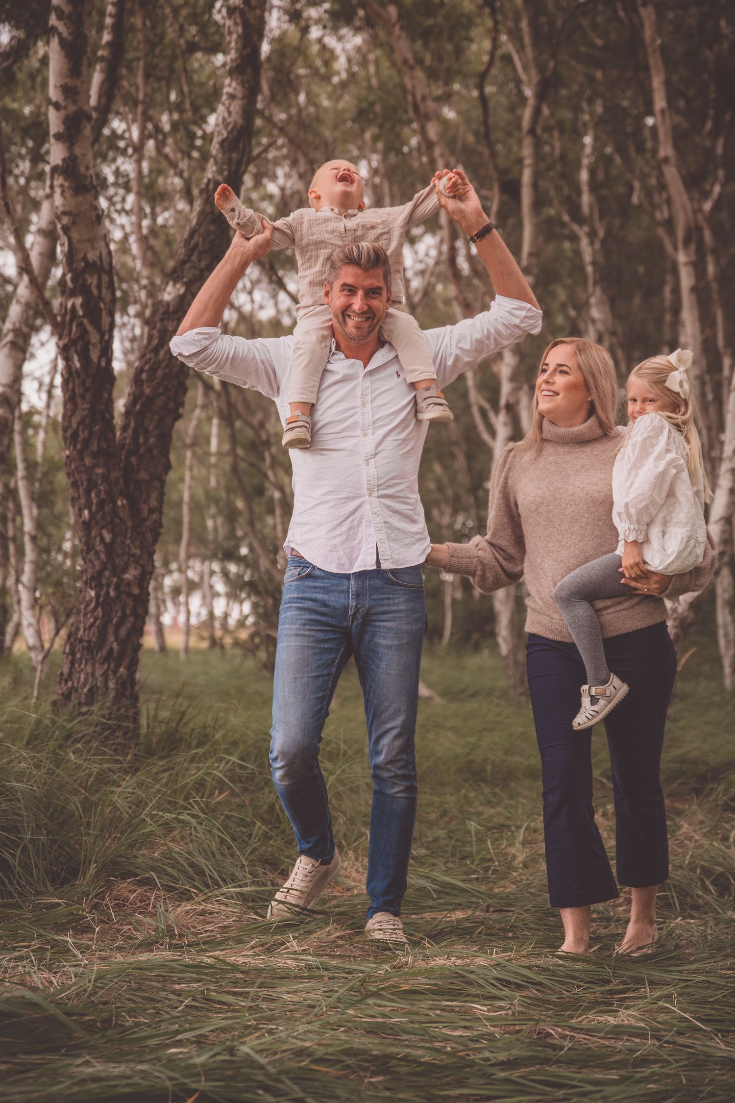A couple in white and beige outfits dance together joyfully in a wooded outdoor setting at sunset.