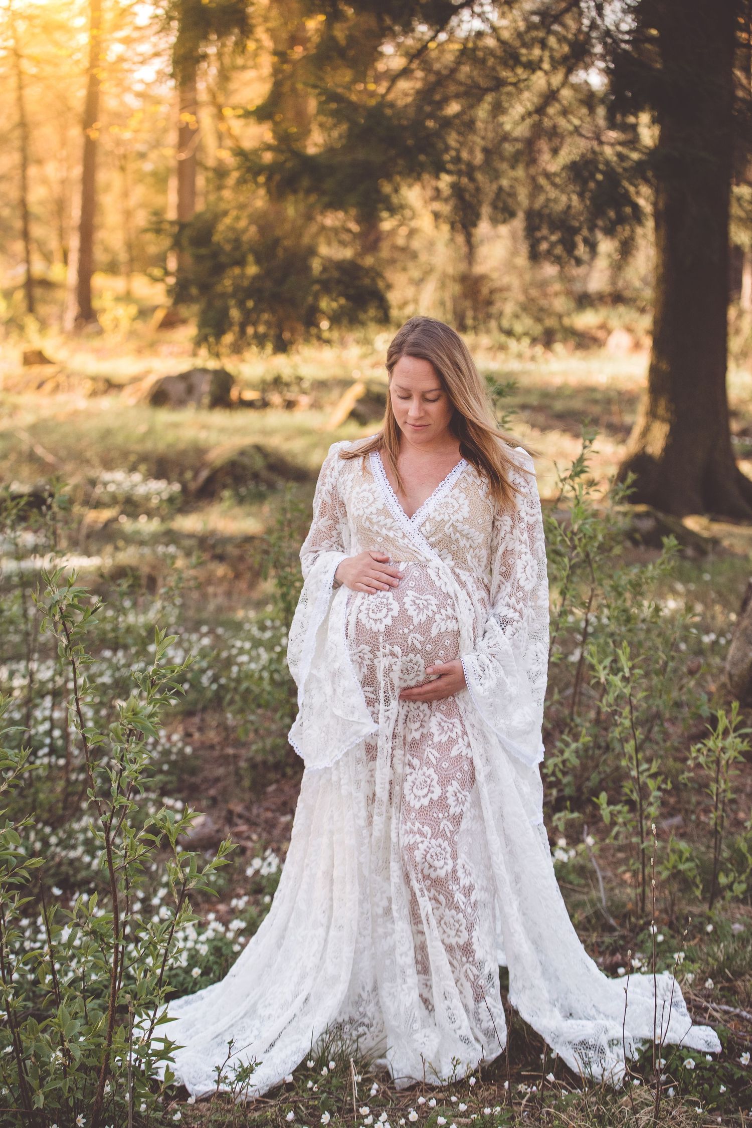 A woman in a flowing white lace dress poses gracefully in a sunlit forest meadow with wildflowers.
