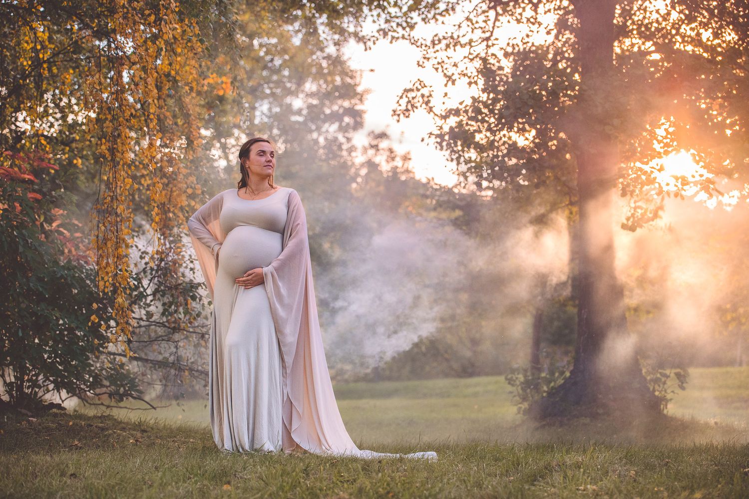 Ethereal maternity photoshoot in a misty park at golden hour with soft sunlight filtering through autumn trees.