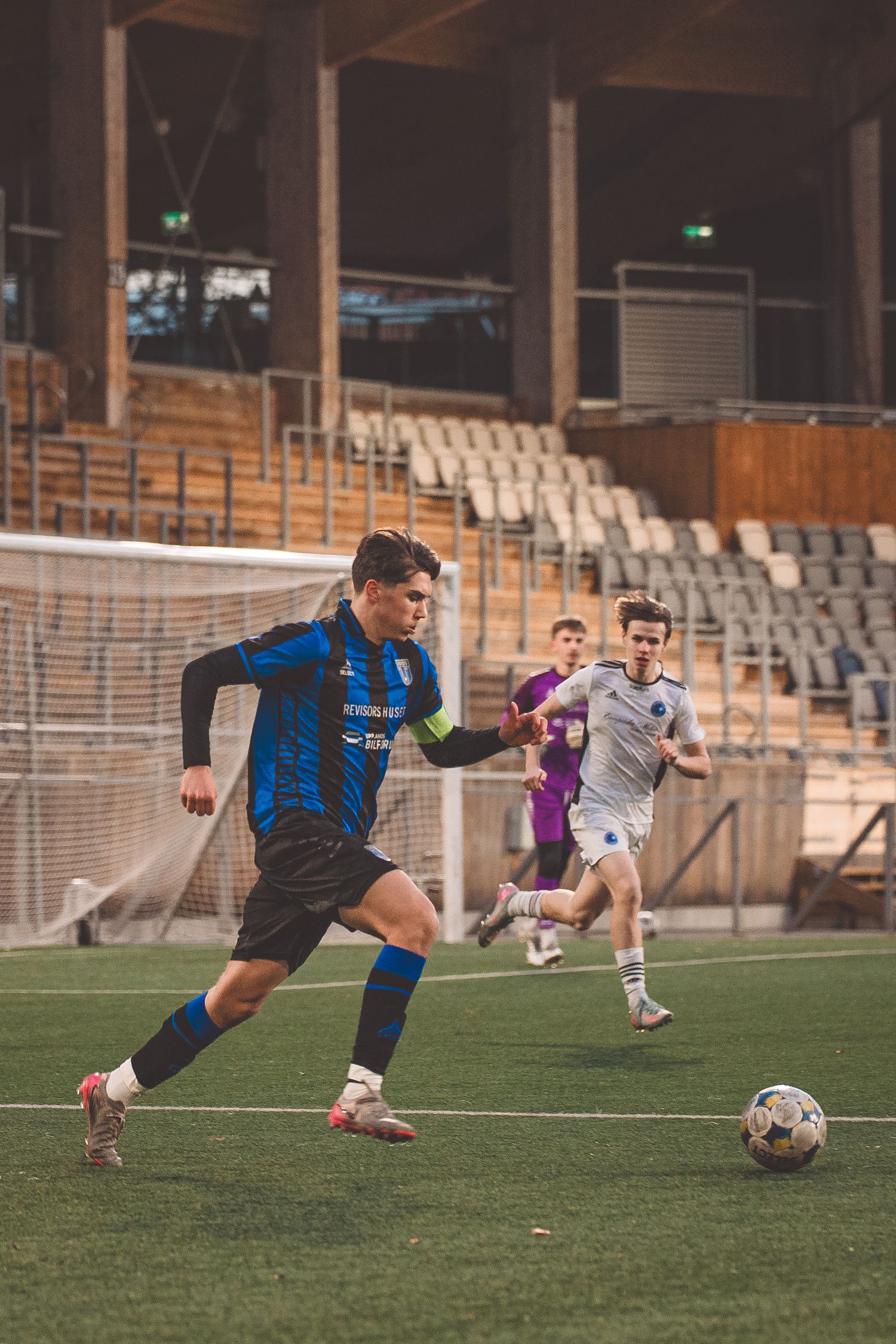 Soccer player in blue and black stripes controls ball during match with opponents approaching.