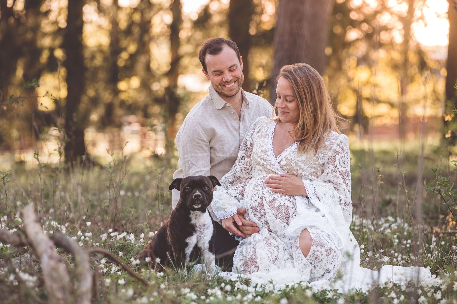 Couple shares intimate moment in field of white wildflowers during sunset with dog nearby.