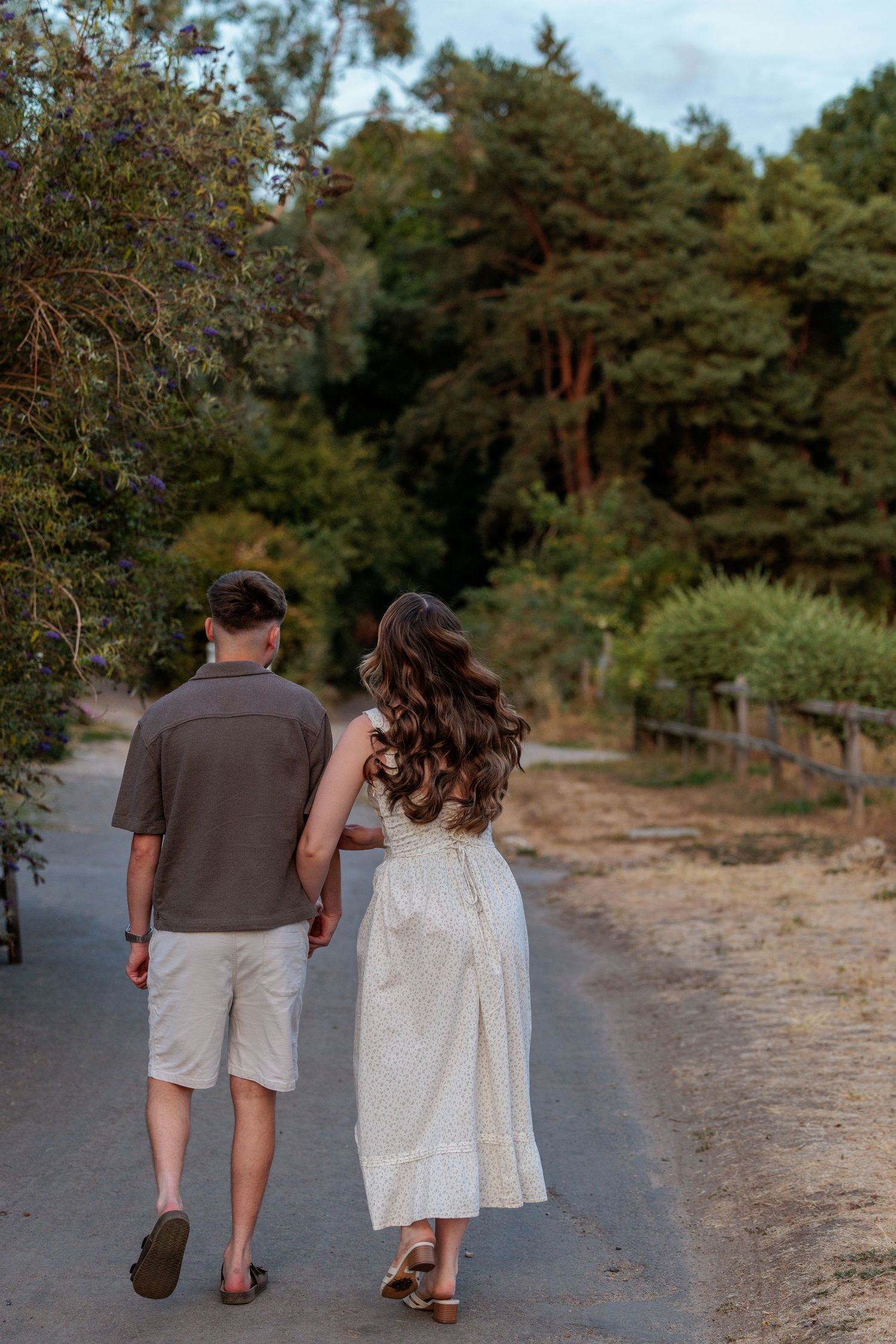 Couple holding hands walking down a tree-lined path, woman in white dress and man in casual shorts.