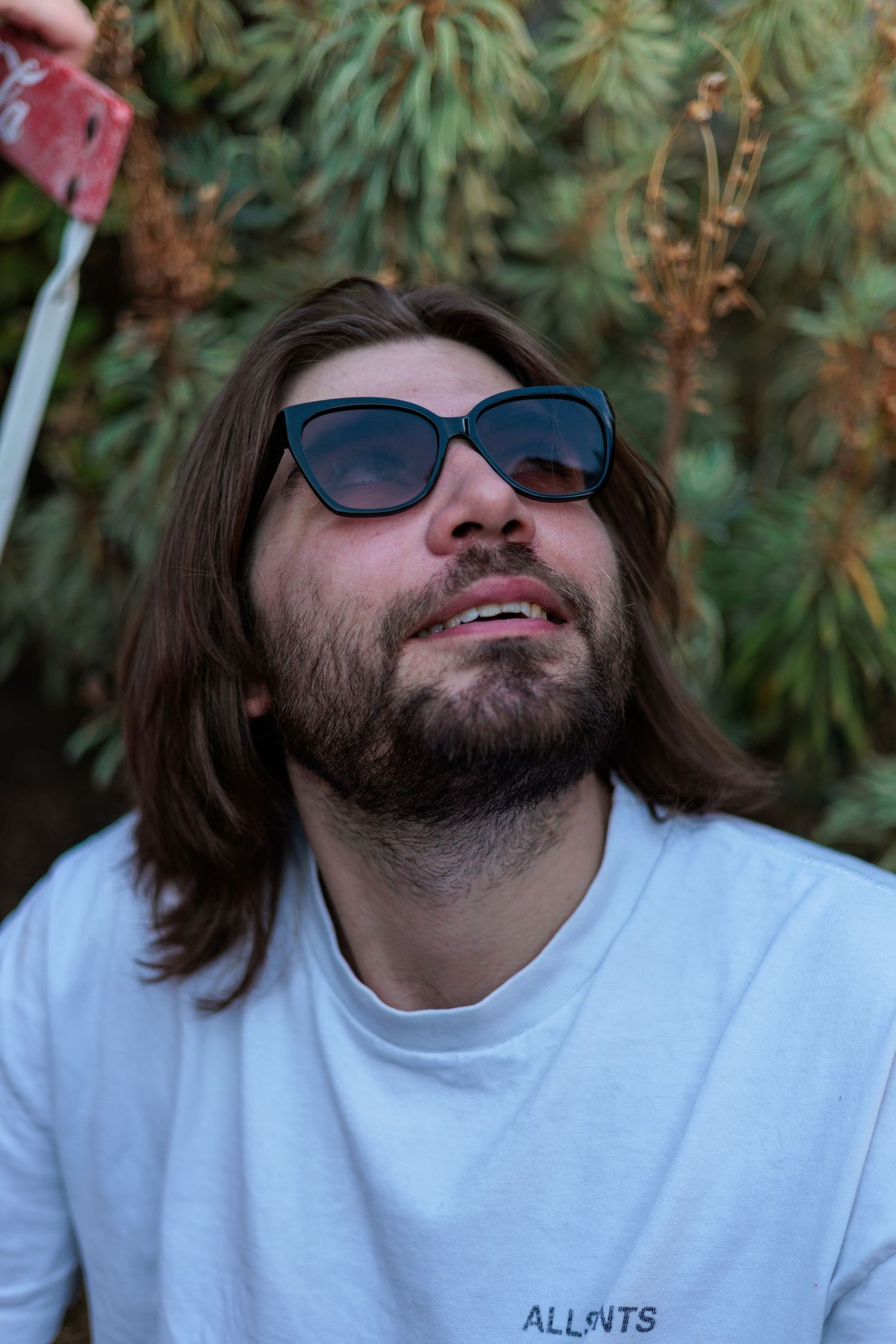 Bearded man with long brown hair wearing blue cat-eye sunglasses and white ALLNTS t-shirt, sitting outdoors near pine trees.