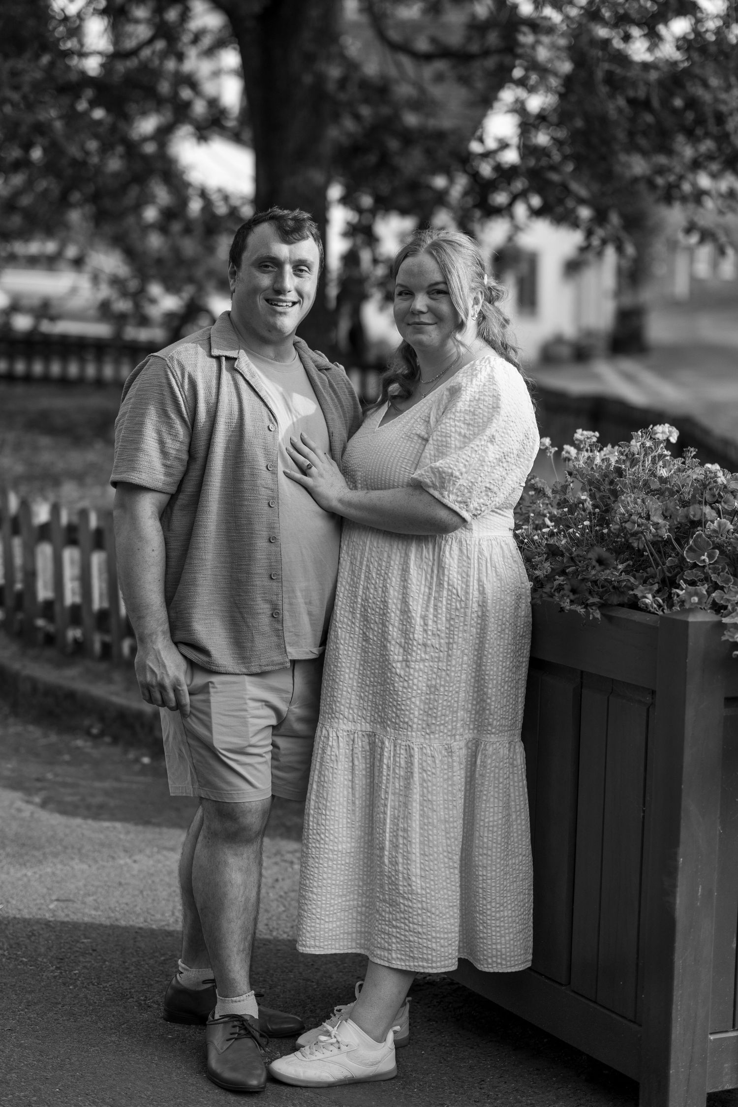 Black and white photo of a smiling couple posing outdoors near a flower planter and white picket fence.