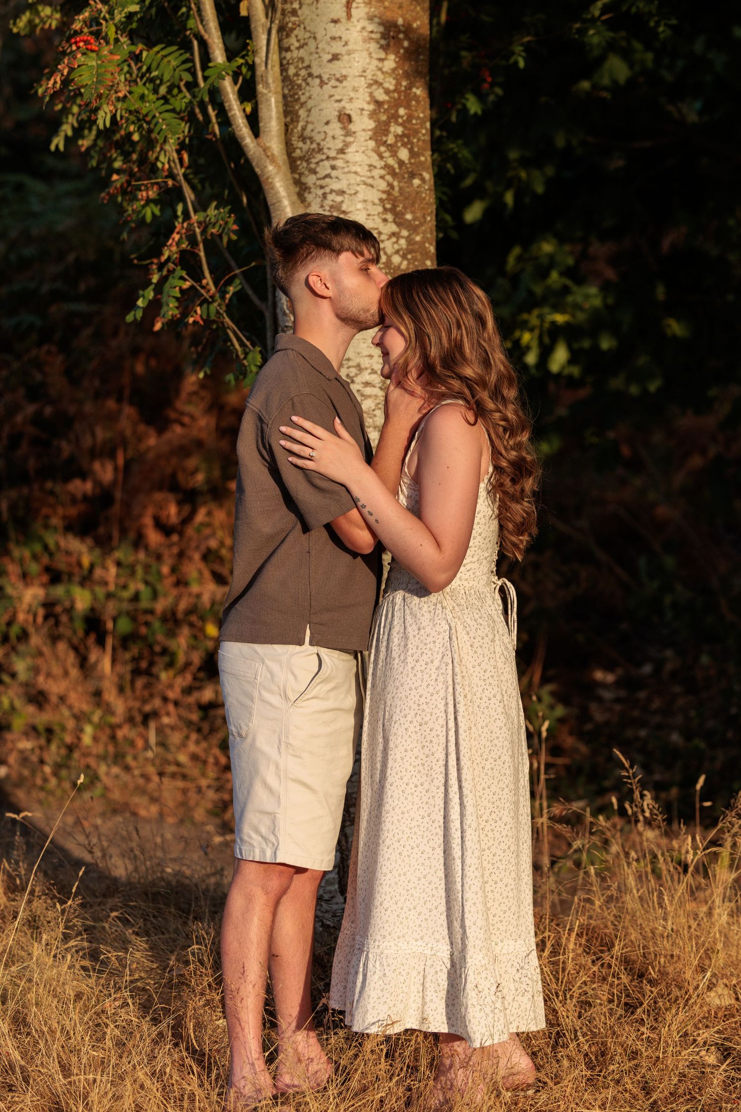 A young couple shares a romantic kiss in golden hour sunlight beside a tree in a lush forest setting.