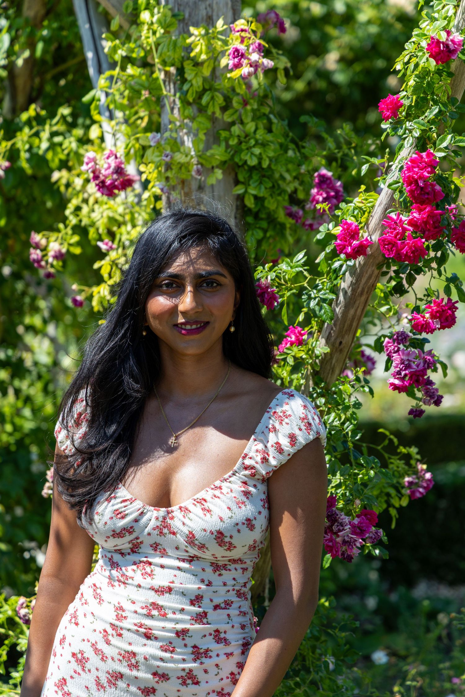 A woman in a floral dress poses in a garden surrounded by vibrant pink climbing roses and lush greenery.