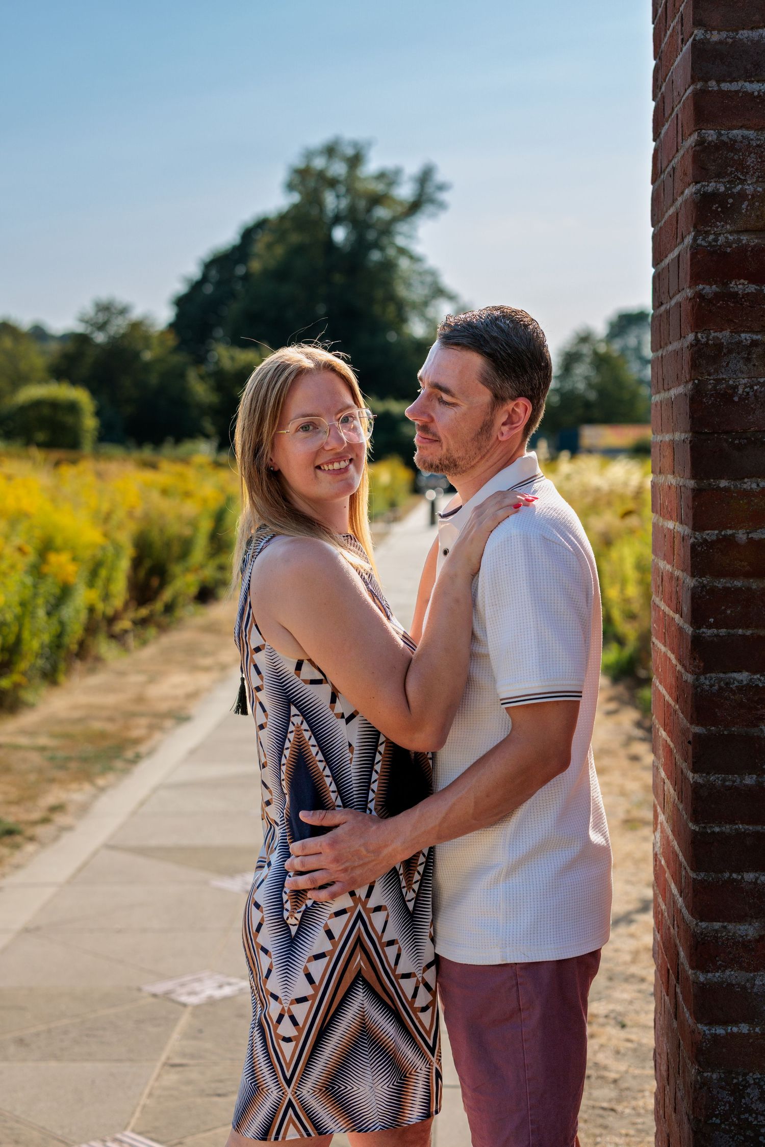 A smiling couple embraces outdoors on a sunny day, with lush greenery and a brick wall in the background.